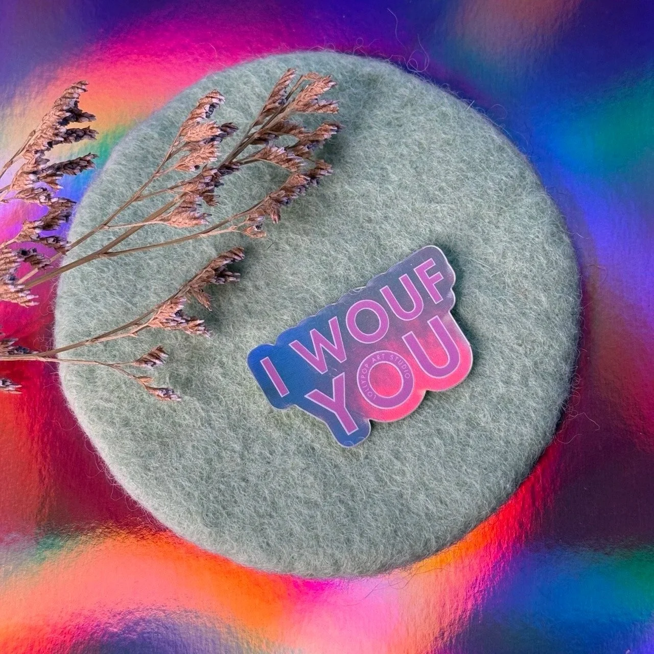 Un tampon en coton gris, décoré d'une étoile en stickers colorés avec le message "I WOUF YOU" et une branche séchée de fleurs violettes, posé sur un fond holographique coloré.
