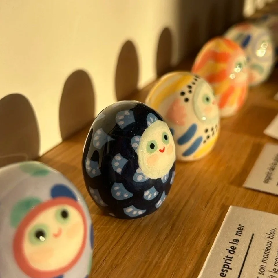 Figurettes en céramique peintes avec des visages de style kawaii et des motifs colorés, exposées sur une étagère en bois.