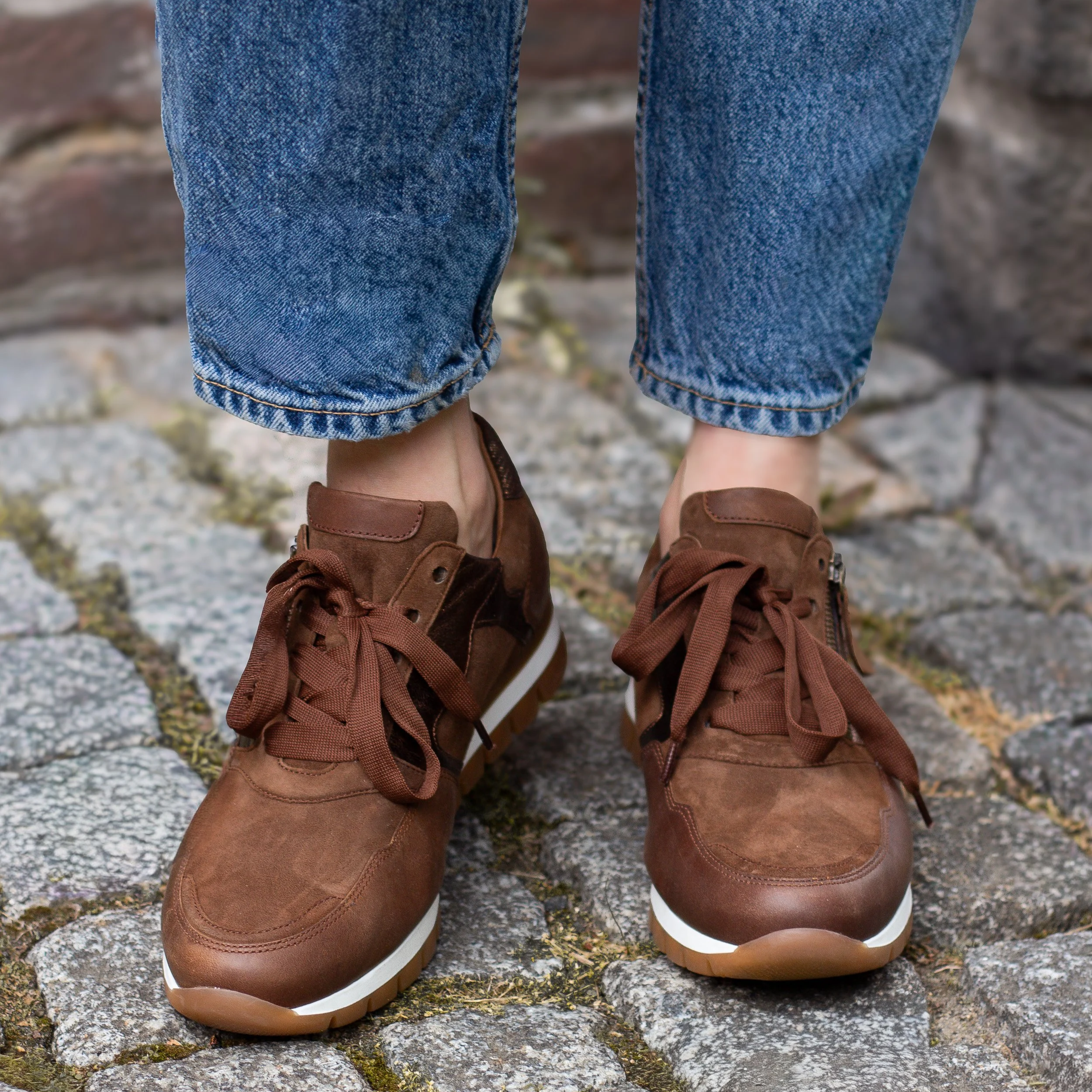 Chaussures de marche brunes portées par une personne portant un jean, sur un chemin pavé en pierre.