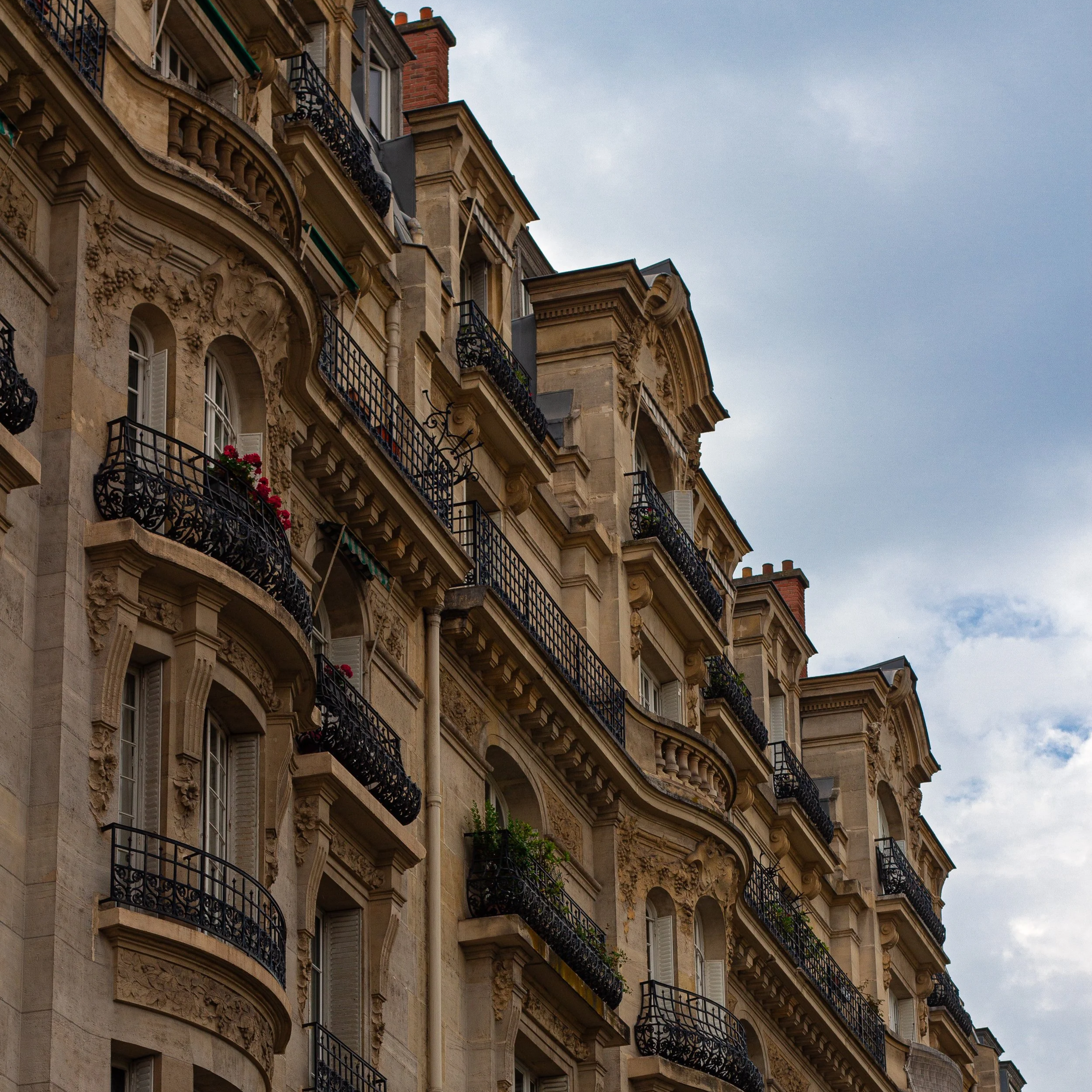 Façade d'un immeuble haussmannien avec balcons en fer forgé et fenêtres ornées de fleurs, sous un ciel nuageux.