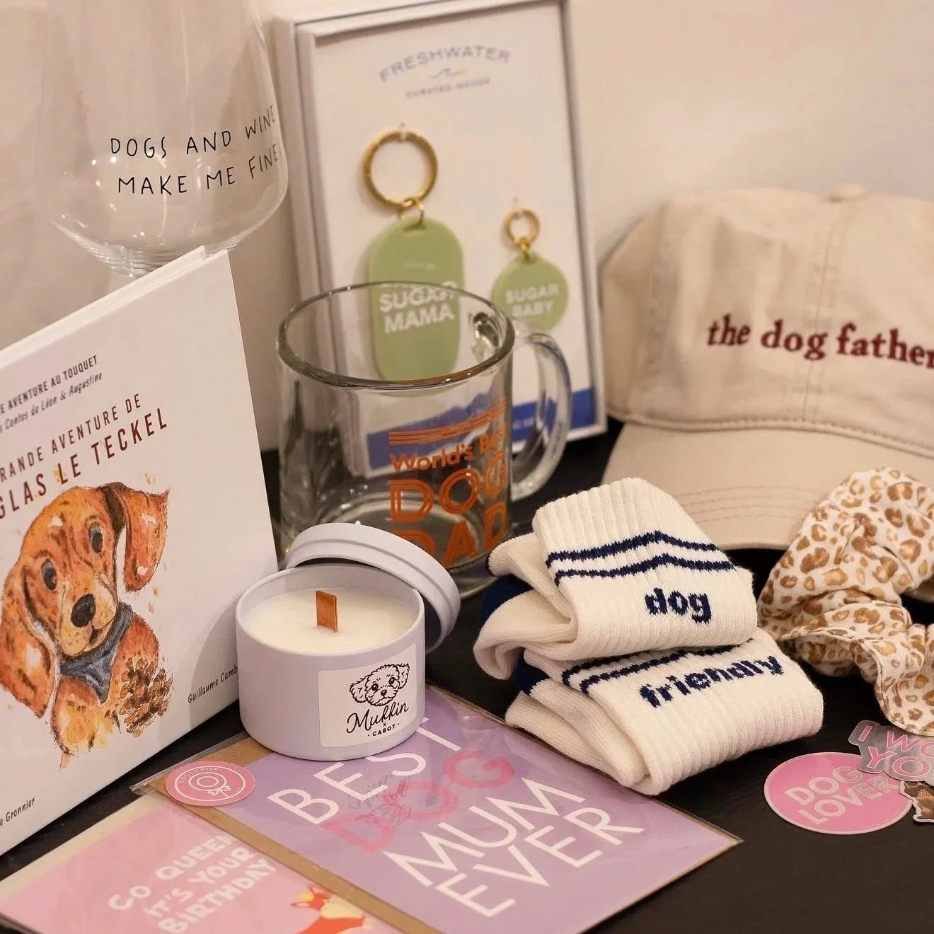 Collection d'objets liés aux chiens, notamment un livre illustré, un mug en verre, une casquette, des chaussettes, une bougie, une écharpe et des stickers. Le thème central est l'amour pour les chiens et la fête des mères.