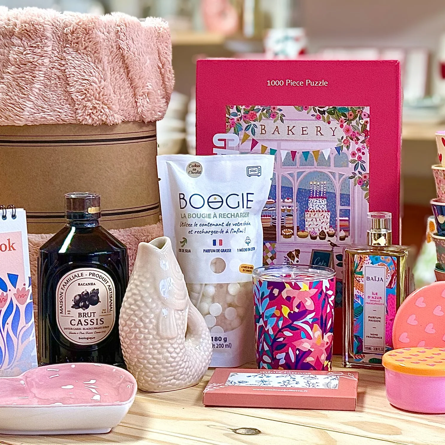 Divers objets de décoration et cadeaux, incluant un puzzle, une bouteille de parfum, une bougie, un vase en forme de poisson, et divers autres articles colorés sur une table en bois.