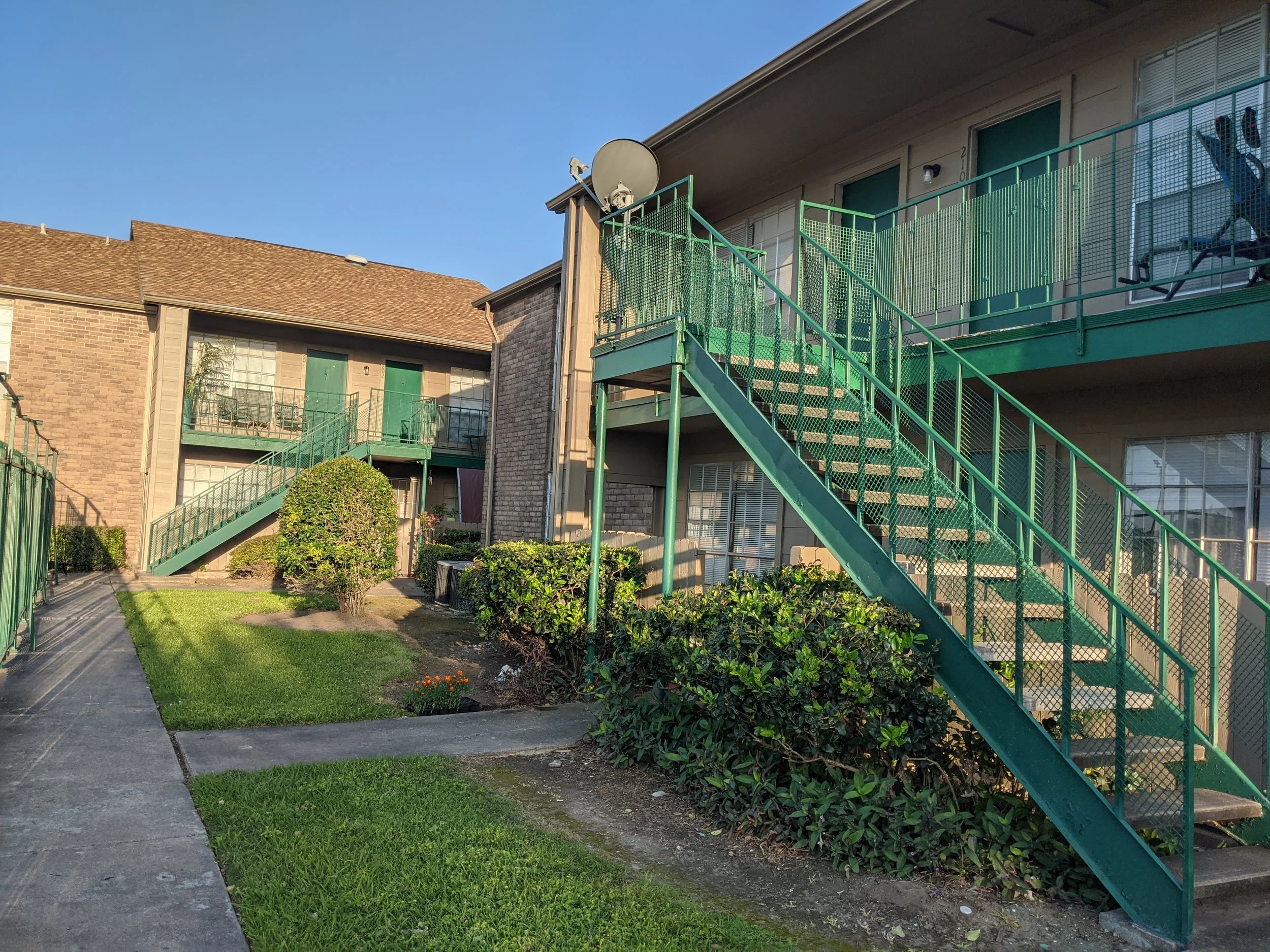 Exterior of a two-story brick apartment building at Greyfield Apartment Homes, with a green metal staircase and balcony railings, ground-floor units with green doors, and landscaped shrubs and flowers along a walkway under a clear blue sky.