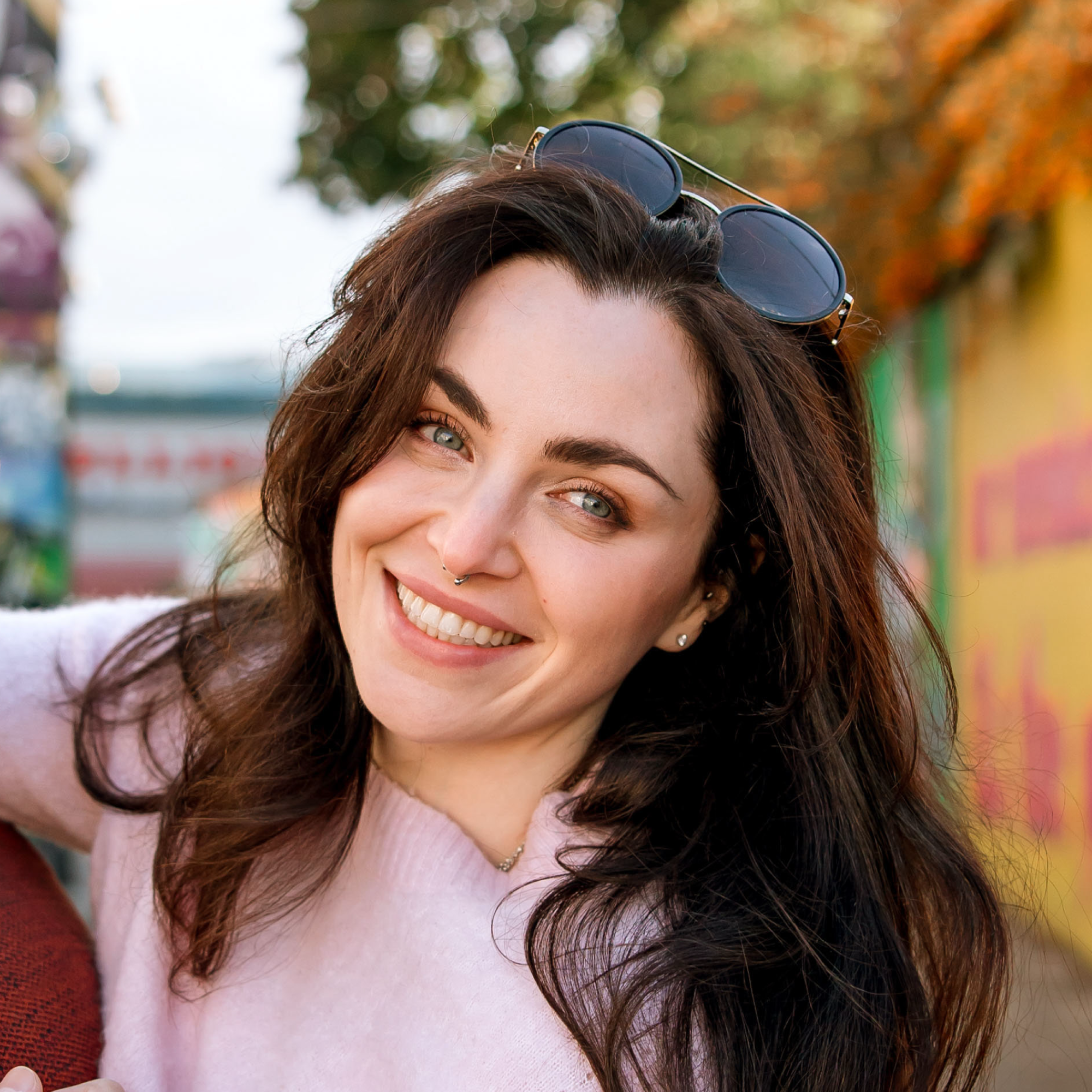 Close-up of smiling woman with brown hair and glasses on her head wearing a pink top, outdoors with autumn-colored trees in background.