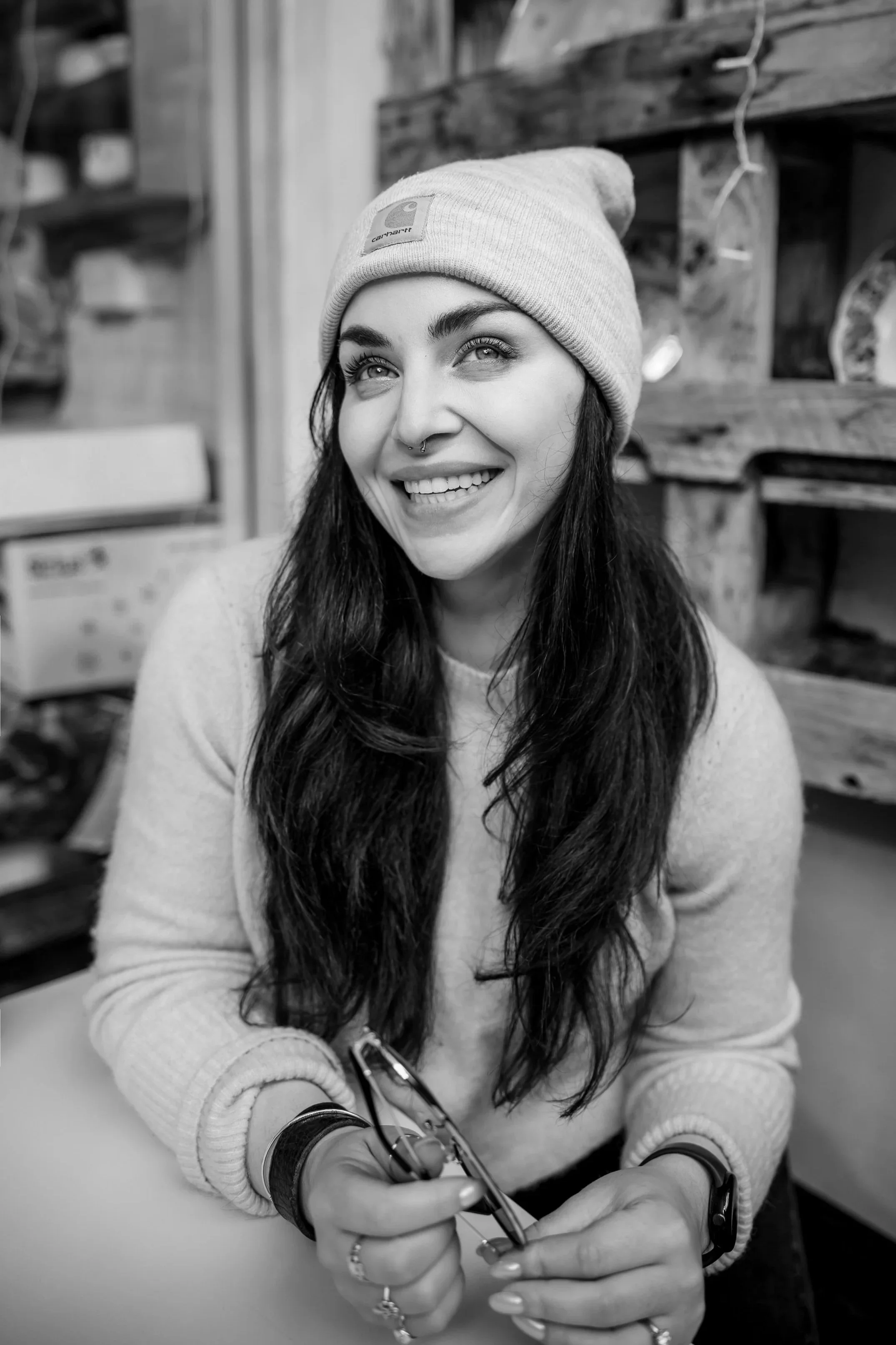 A woman with long dark hair wearing a beanie with a logo, smiling and looking upward, sitting at a table, holding a pair of glasses.