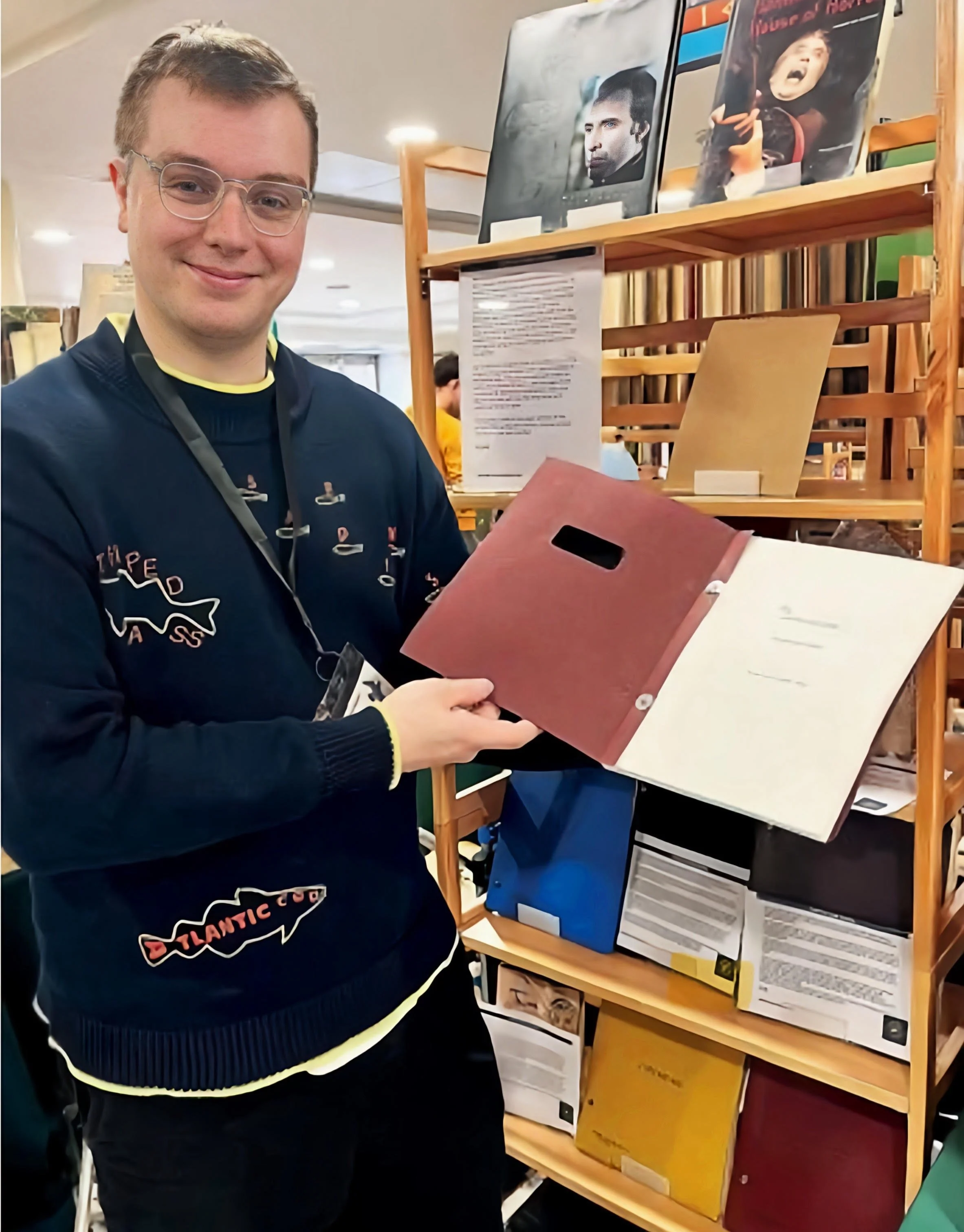 A young man with glasses and a navy sweater with a fish design, holding an open folder, standing in front of a wooden shelf filled with books and artwork.
