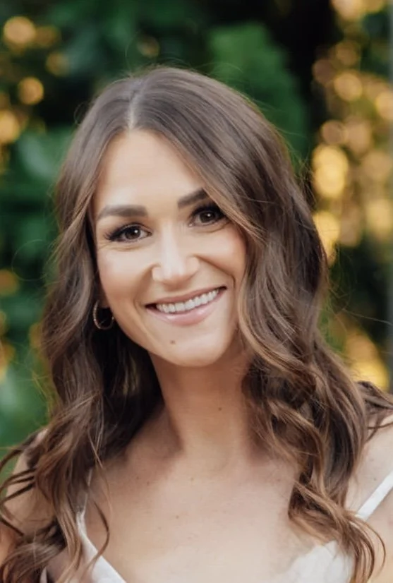 A woman with long wavy brown hair smiling outdoors with greenery and warm bokeh lights in the background.