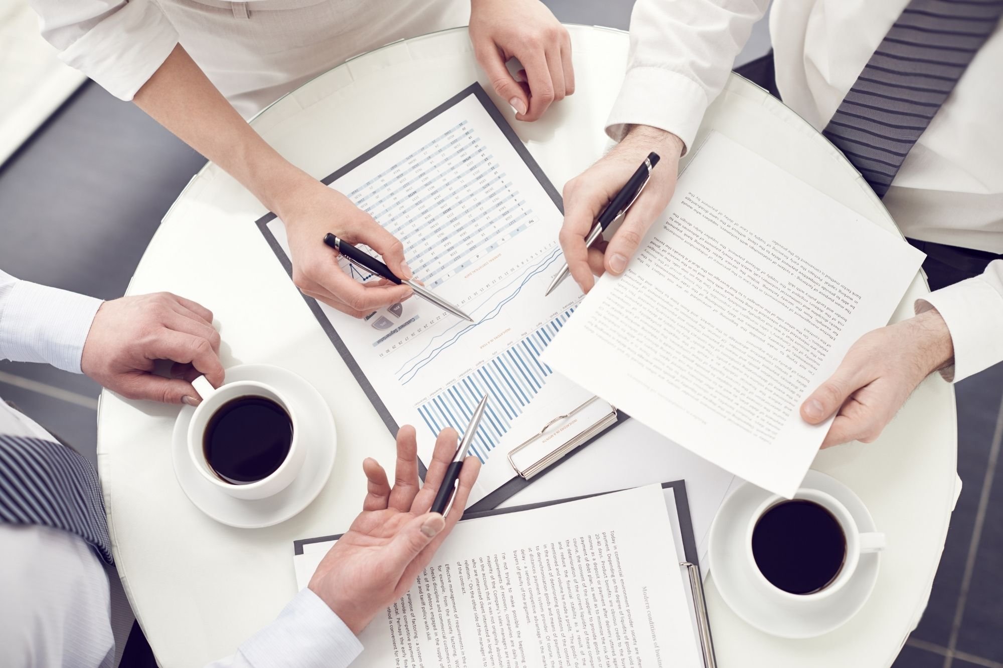 Businessmeeting with three people analyzing financial data and graphs, sitting around a round table with documents, pens, and cups of coffee.
