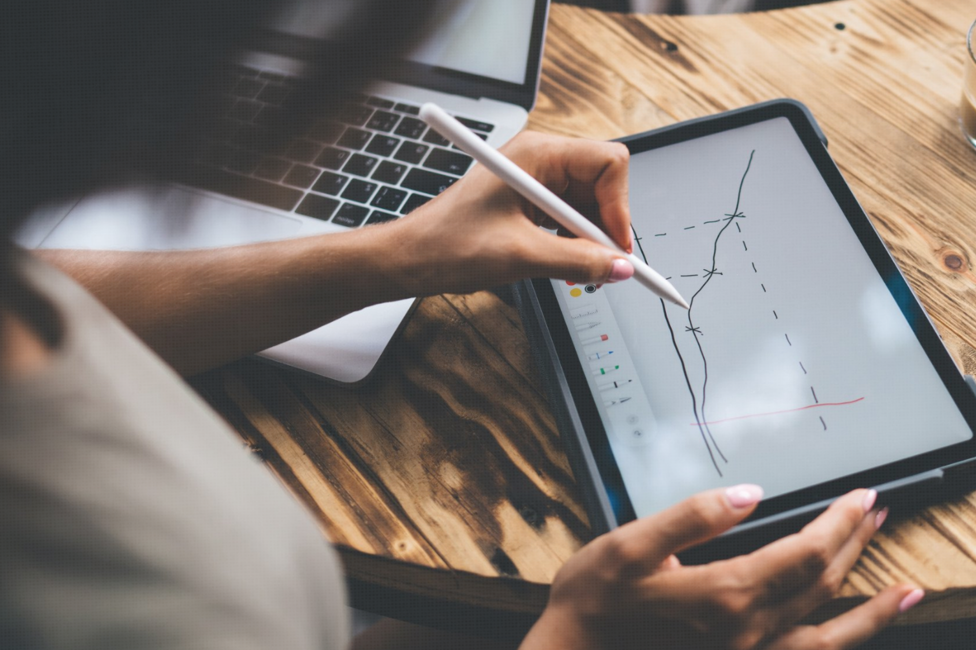 Person drawing graphs on a digital tablet with a stylus. A laptop is visible in the background on a wooden table.