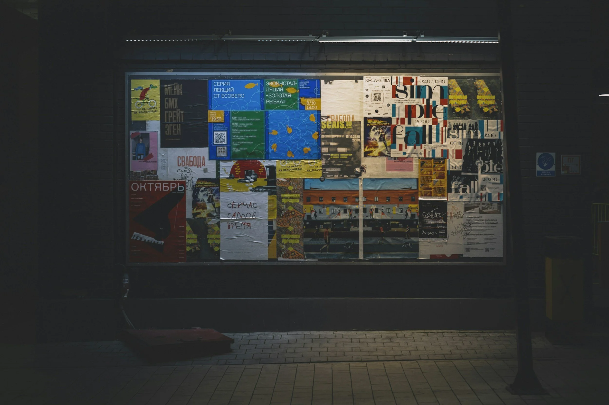 A brightly lit bulletin board covered with various posters, flyers, and notices in different colors and styles, located outdoors at night.