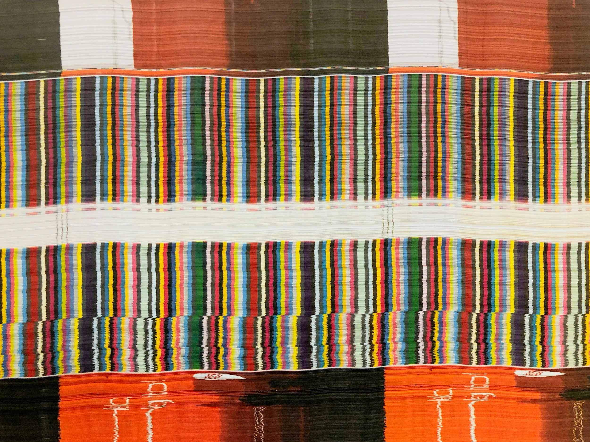 Close-up of a colorful woven textile with horizontal stripes in various bright colors, featuring a section with vertical multicolored stripes.