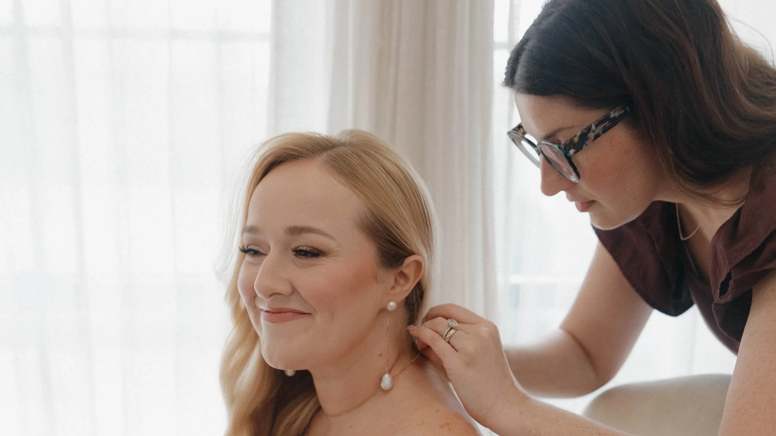 A woman in glasses helps another woman put on a pearl necklace in a bright room with sheer white curtains.