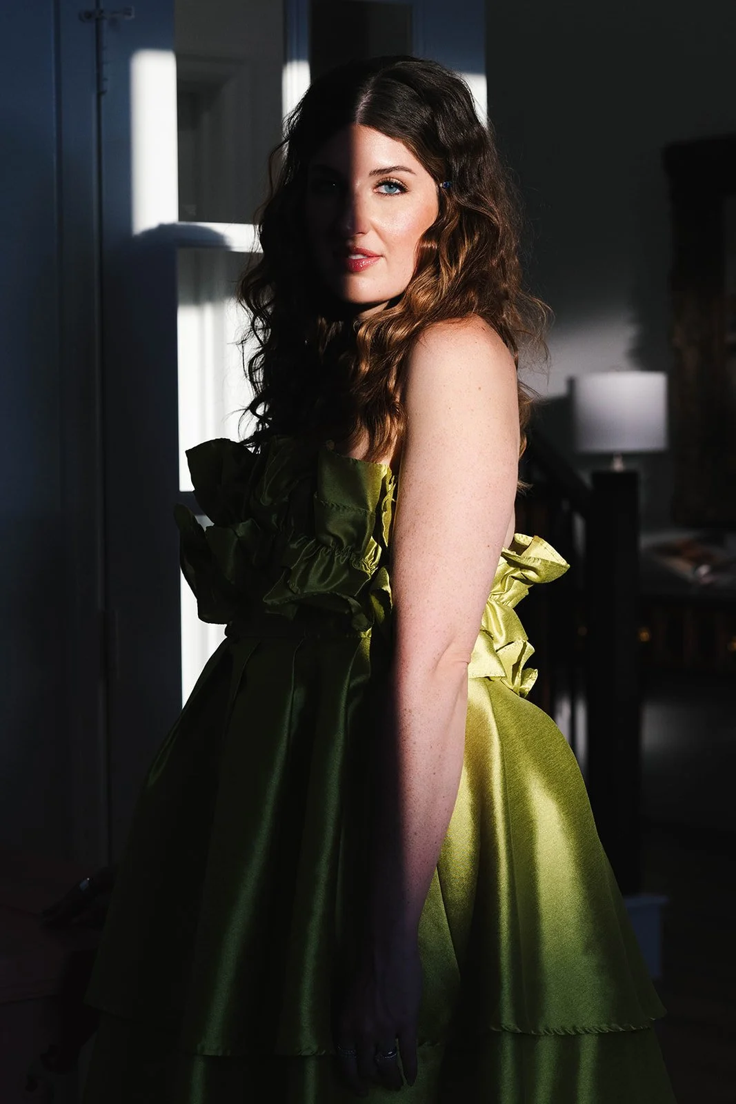 A woman with red curly hair and blue eyes standing in a room with sunlight casting shadows on her face and body, wearing an olive green, ruffled dress.