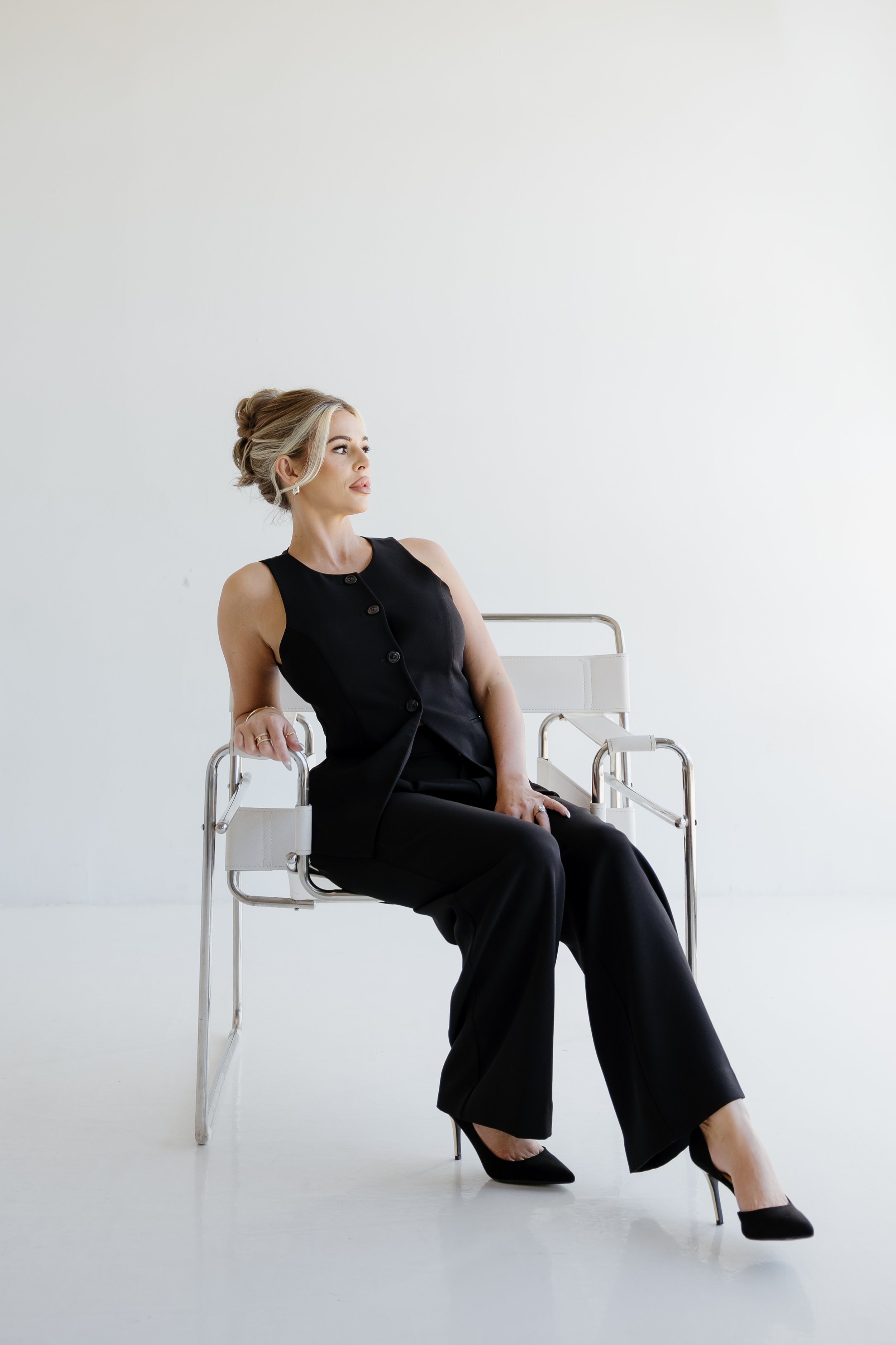A woman in black professional attire sitting on a modern metal and white chair against a plain white background, looking to her left.