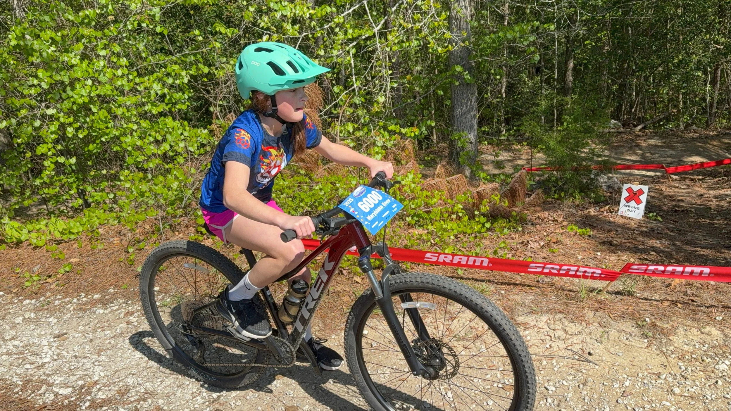 A young girl riding a mountain bike on a trail in the forest during a race, wearing a turquoise helmet, a blue shirt with a racing design, pink shorts, and white socks, with a race plate number 6000 attached to the front of her bike.