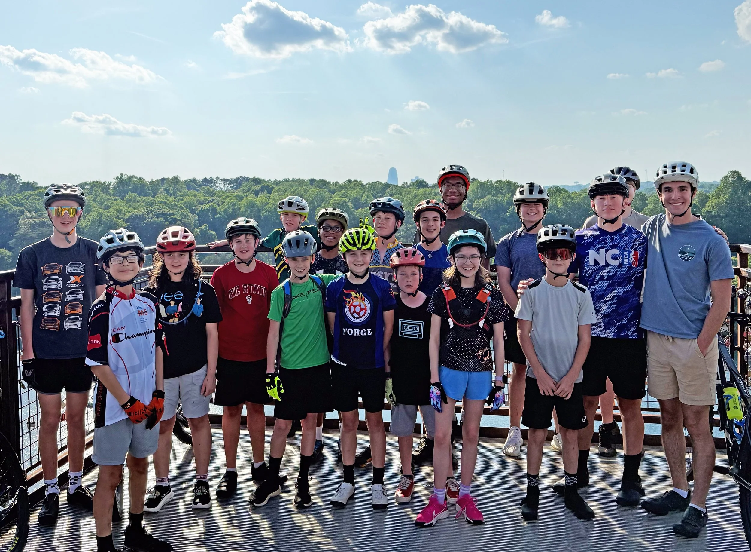 A group of children and a few adults wearing helmets and casual clothes, standing on a viewing platform together outdoors with trees and a skyline in the background.
