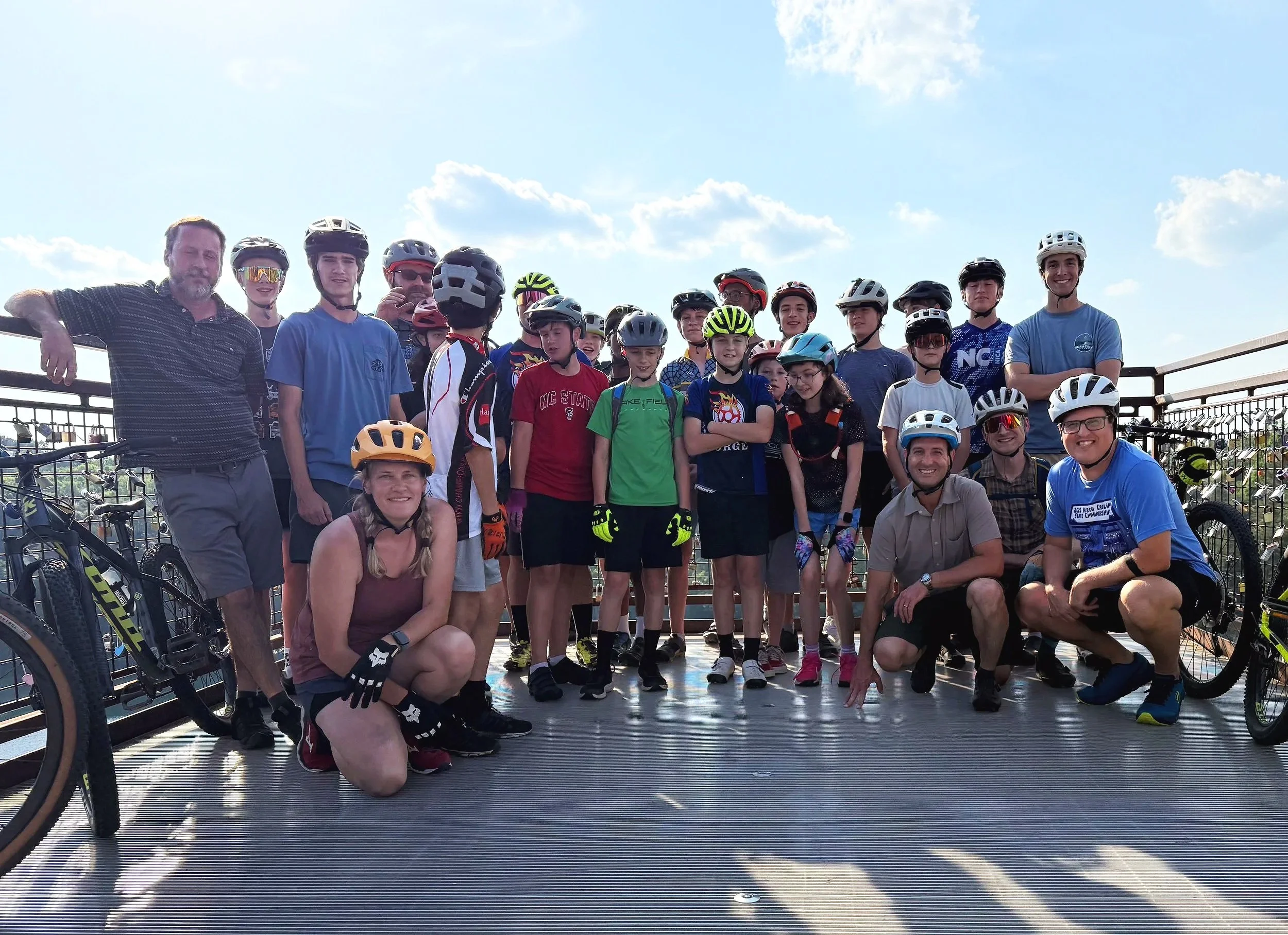 Group of children and adults in biking gear on a bridge, with bicycles and a city view in the background.