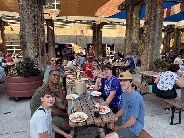 Group of young boys and some adults sitting at a long outdoor wooden table with plates, drinks, and pizza at a casual restaurant or cafe.