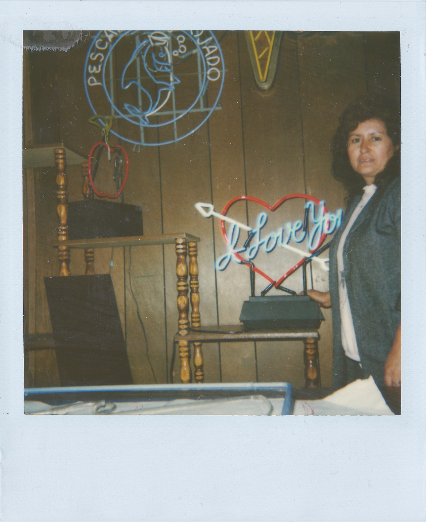 A woman standing next to neon signs, one says 'I Love You' inside a red heart and another says 'Pescados y Mariscos' with a fish outline. The setting appears to be a restaurant or bar with wooden paneling.