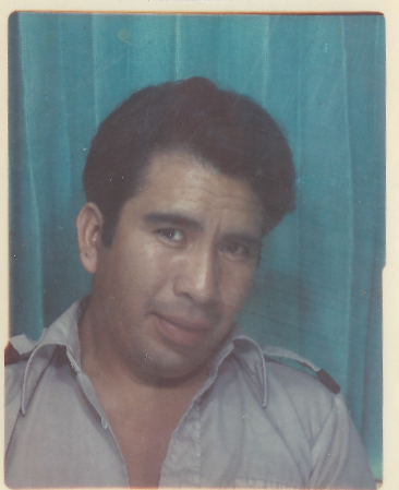 A man with dark hair in a light uniform shirt, standing in front of a teal curtain, looking at the camera.