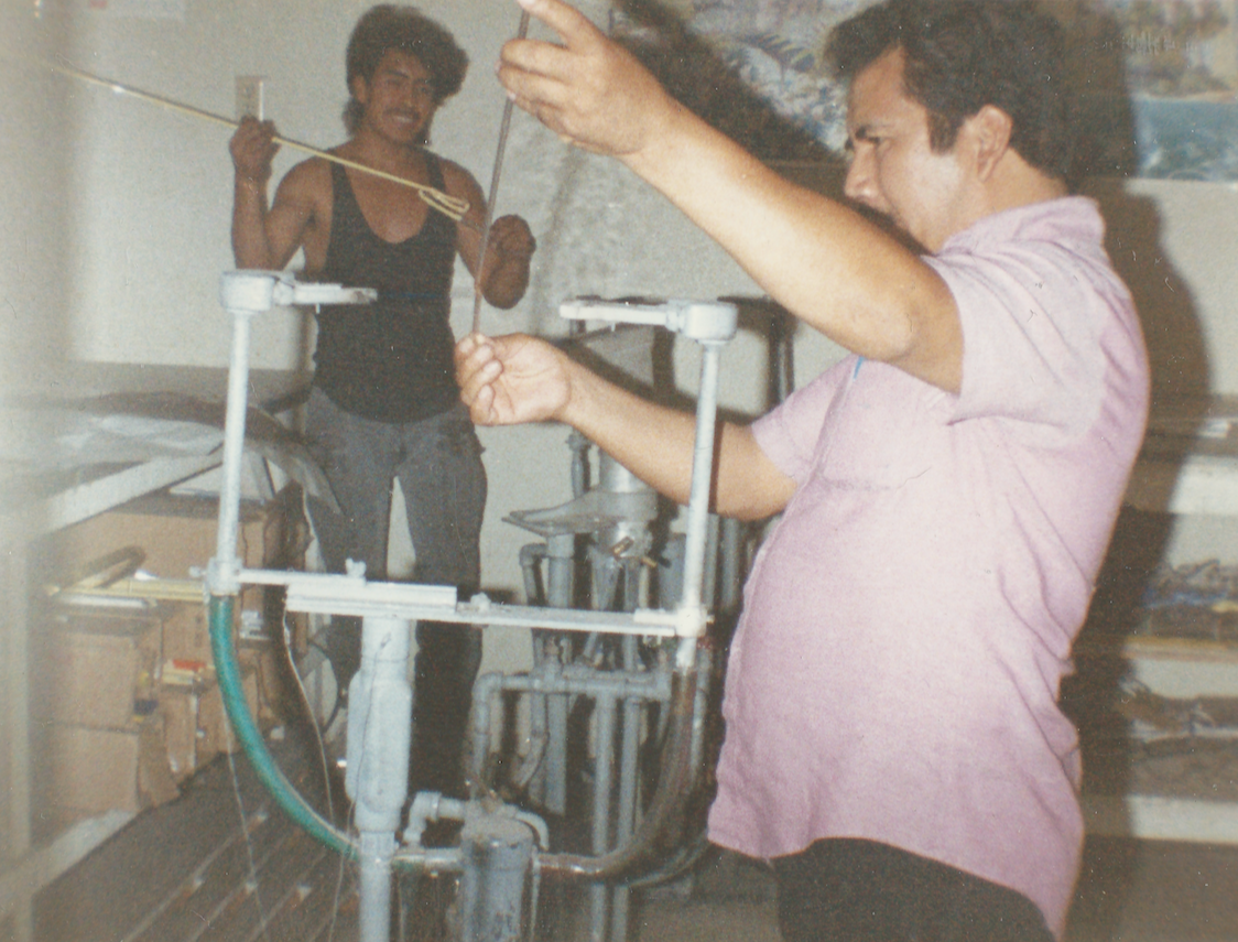 Two men in a workshop, one appears to be working on a metallic apparatus while the other stands nearby holding a yellow string. The man working on the apparatus is wearing a pink shirt, and the other man is in a sleeveless black shirt. There are tools and boxes on shelves around them.