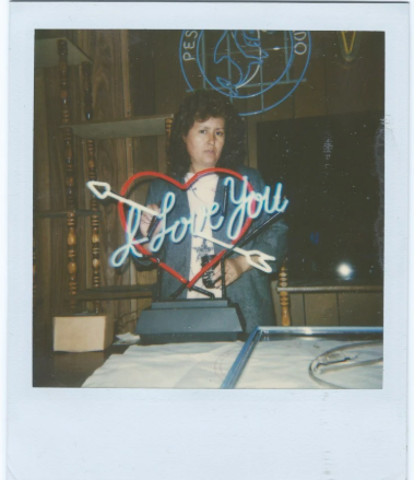 A woman standing behind a table with a neon sign that says 'Love You' inside a heart, with arrow decorations, and a wooden background.