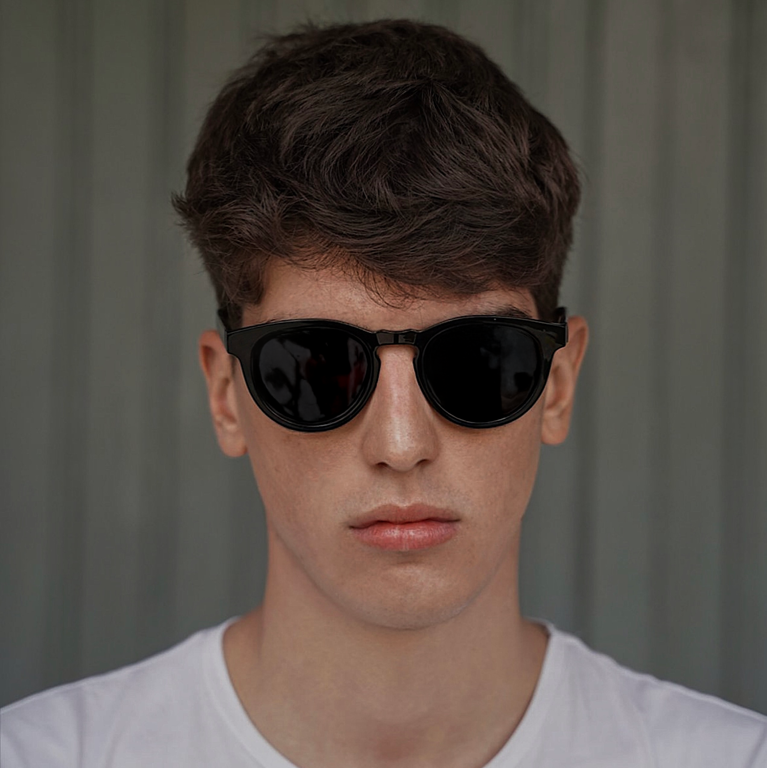 A young man wearing black sunglasses and a white shirt, standing in front of a gray background.