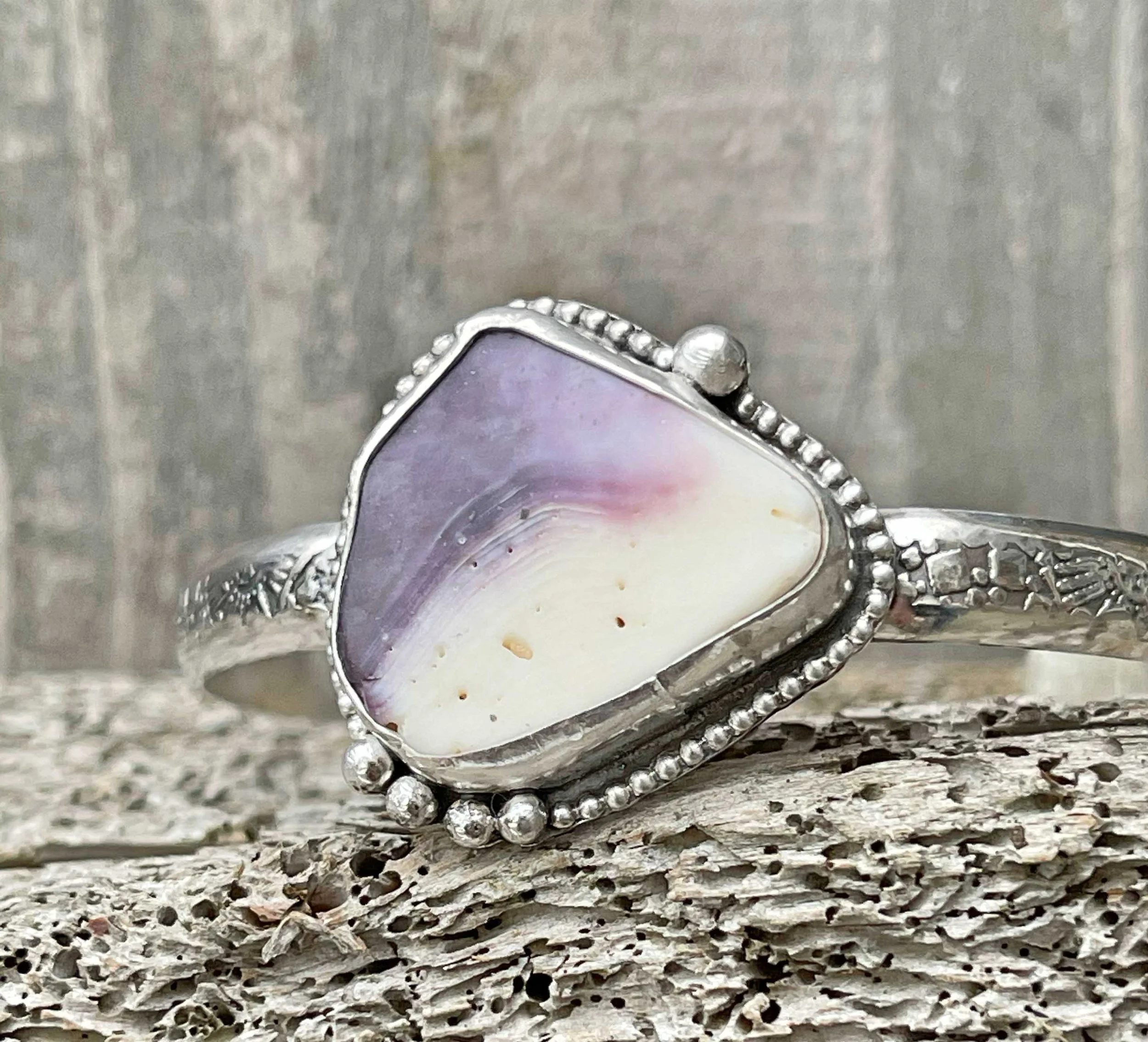 Silver ring featuring a large, triangular shell with purple and white coloring, decorated with small silver beads around the shell and on the band, resting on a textured, weathered surface with a wooden background.