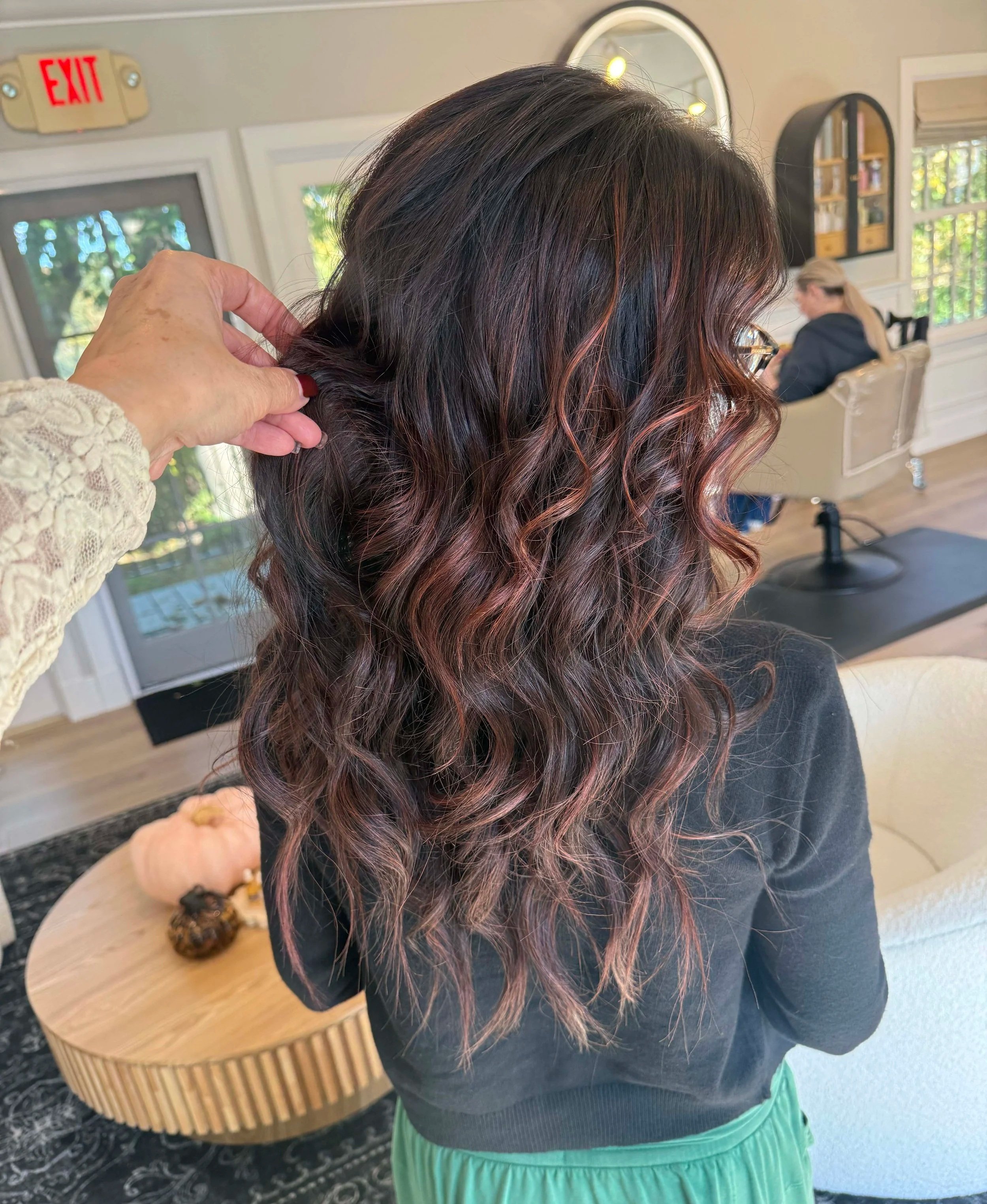 Close-up of a person with long, curly, dark brown hair with reddish highlights. Someone is gently holding a section of the hair. The background shows a cozy indoor setting with a woman sitting on a cream-colored sofa, a window, and decorative wall mirrors.