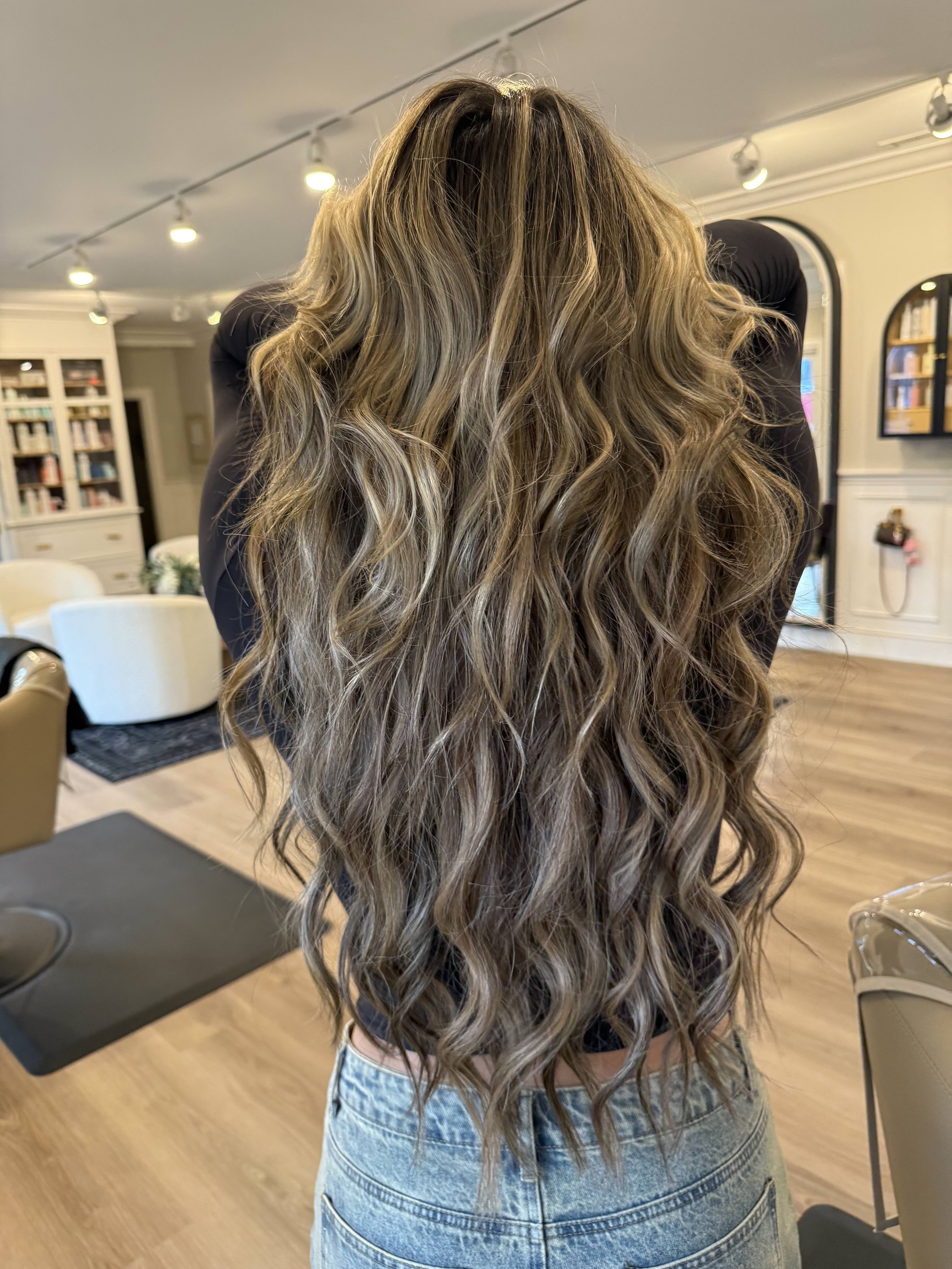 A woman with long, wavy, light brown hair standing in a cozy, well-lit room with wooden flooring and white furniture.