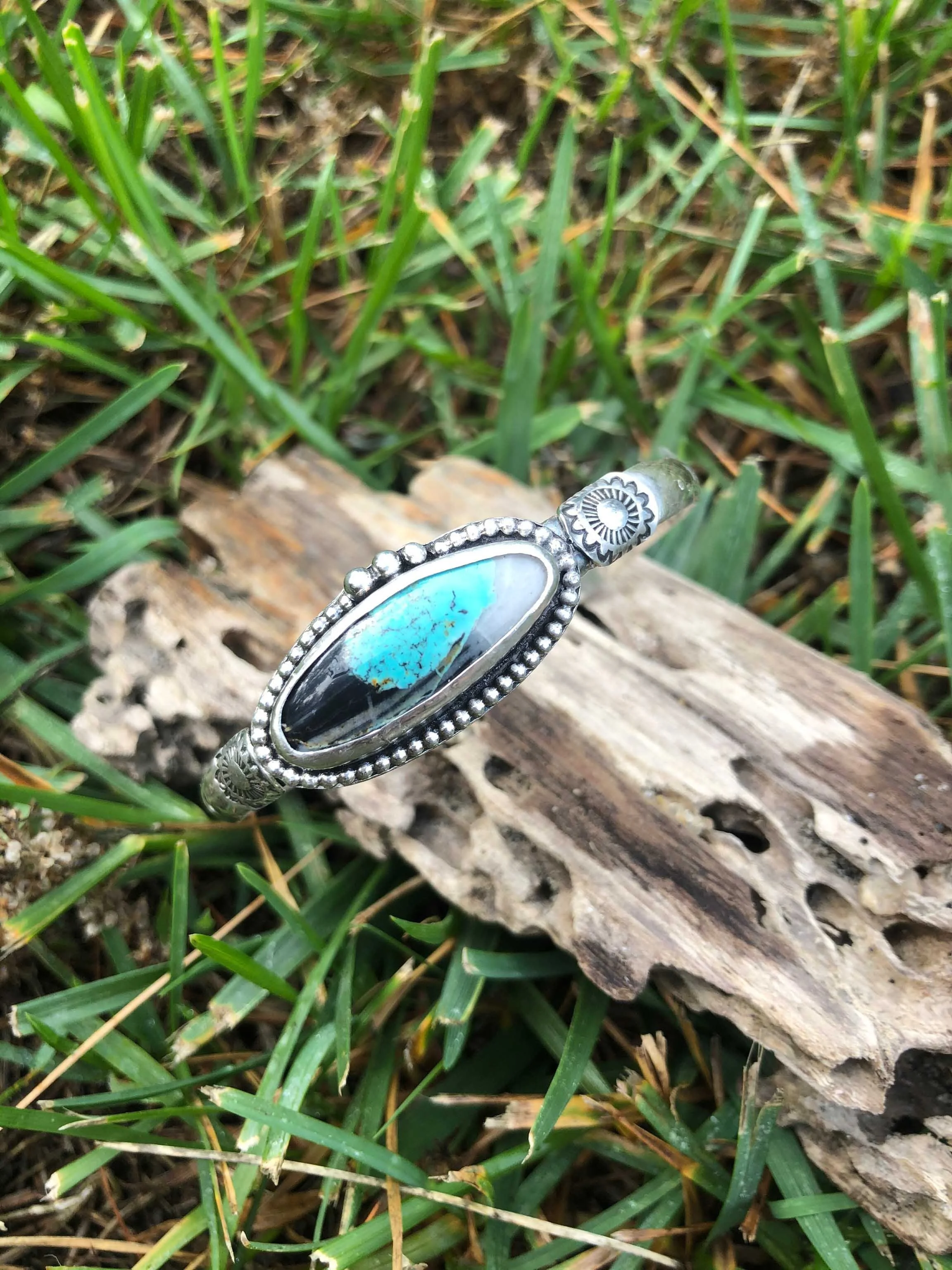 Silver bracelet with a large oval turquoise and black stone, resting on a piece of driftwood in grass.