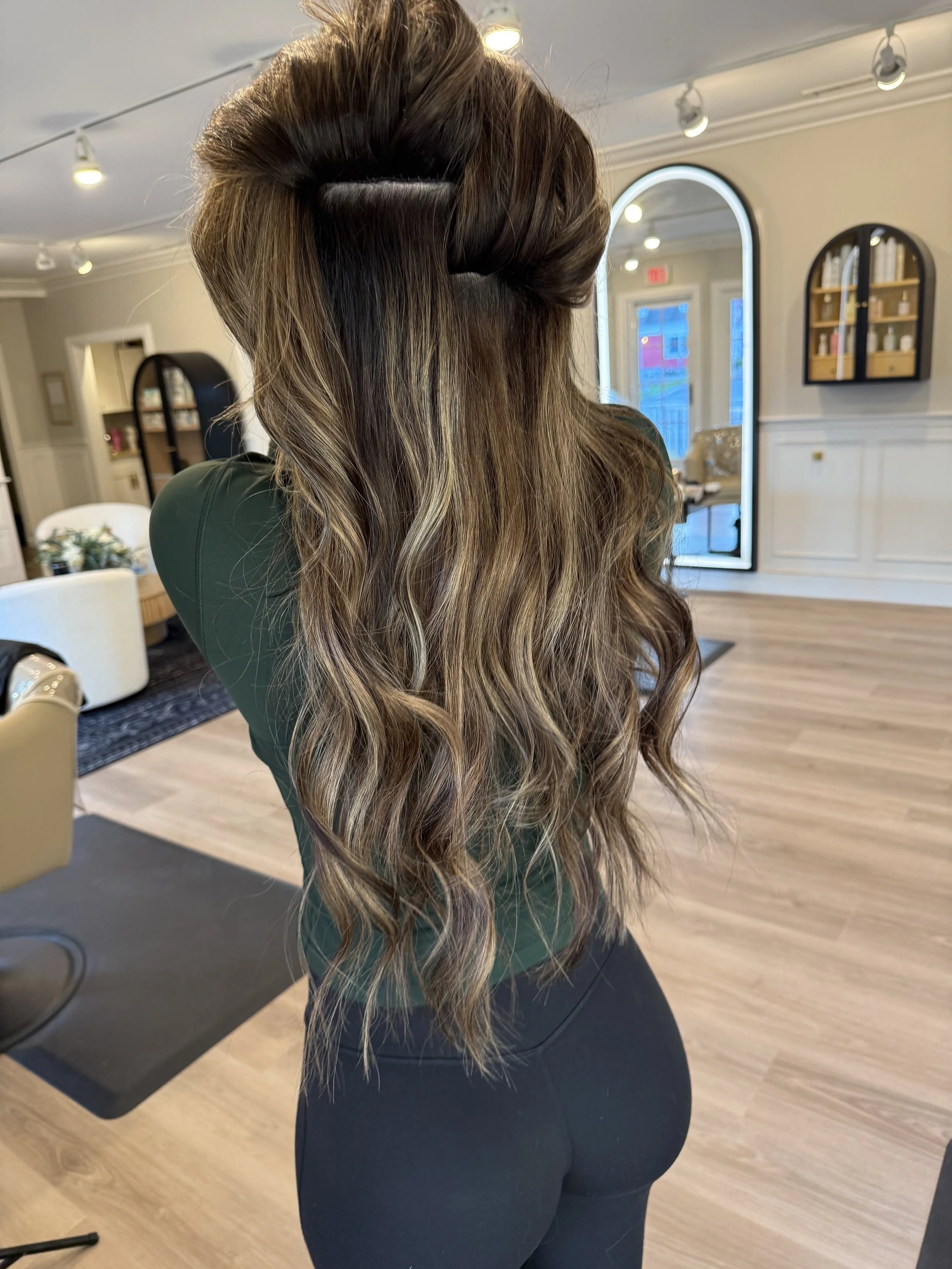 A woman with long, styled wavy brown hair with lighter highlights, seen from the back, in a hair salon with large arched windows and white walls.
