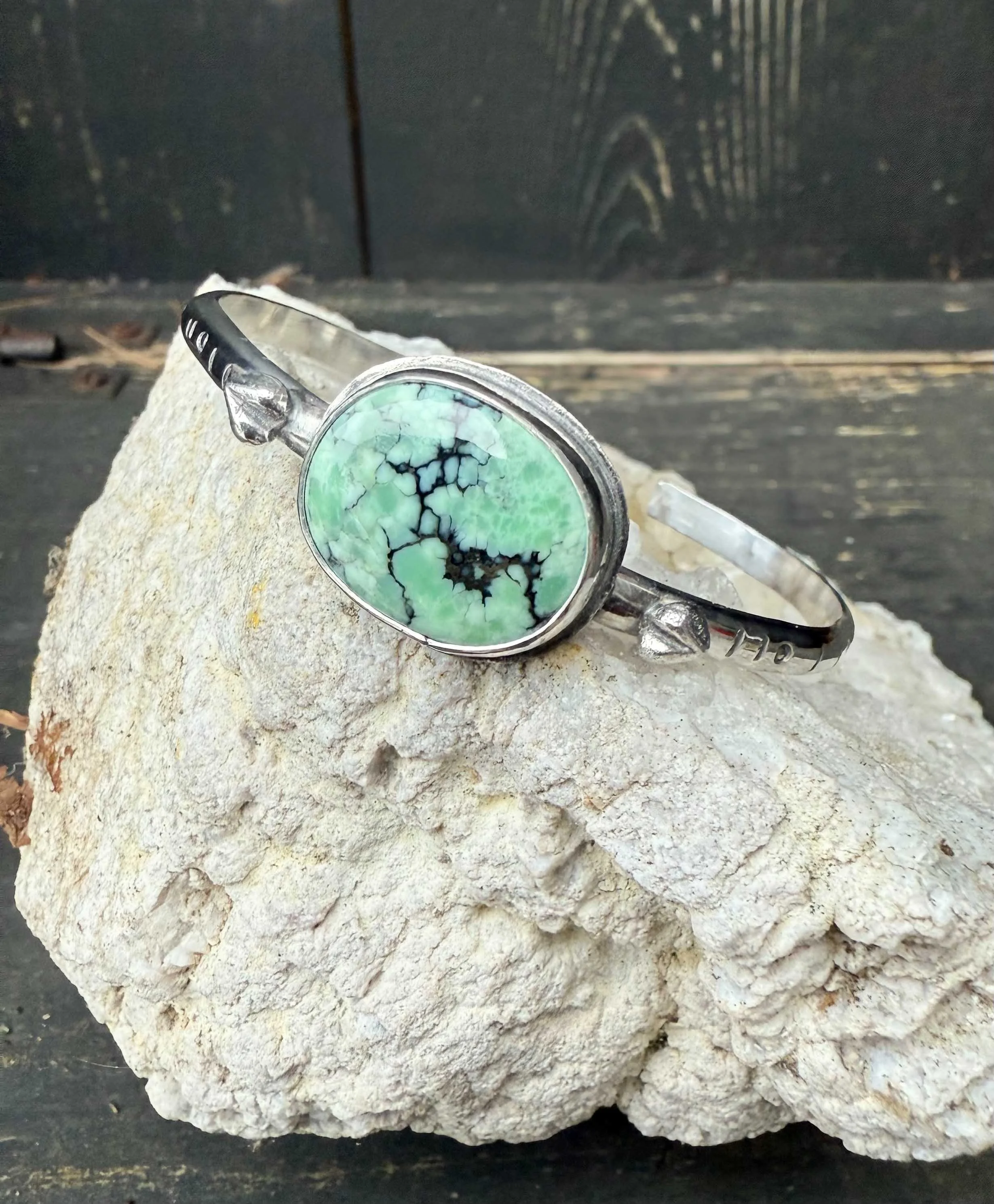 Silver bracelet with a green and black marble-like stone, resting on a large beige rock.