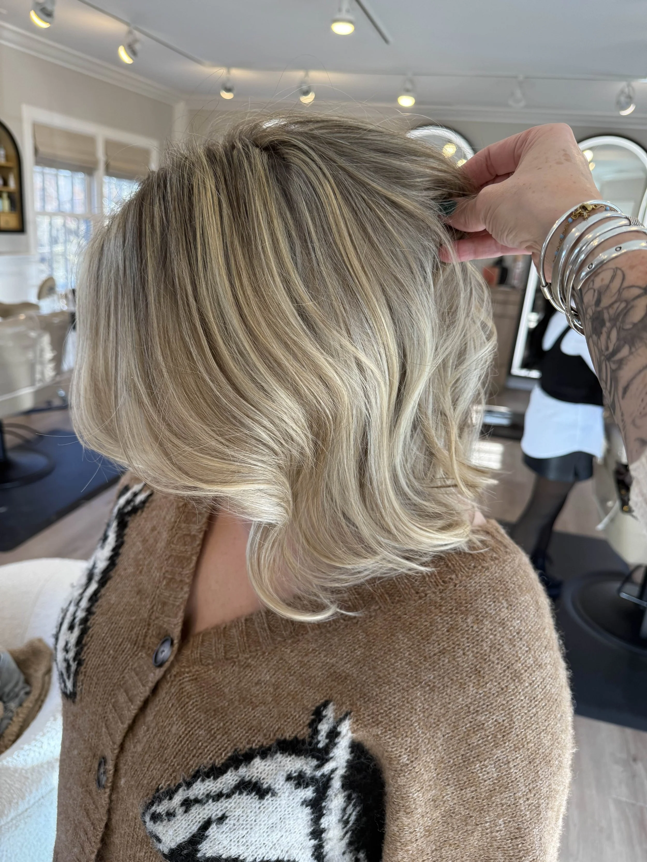Close-up of a woman with shoulder-length blond hair styled in loose waves inside a salon, with a hairstylist's hand adjusting her hair; salon interior with mirrors and lighting.