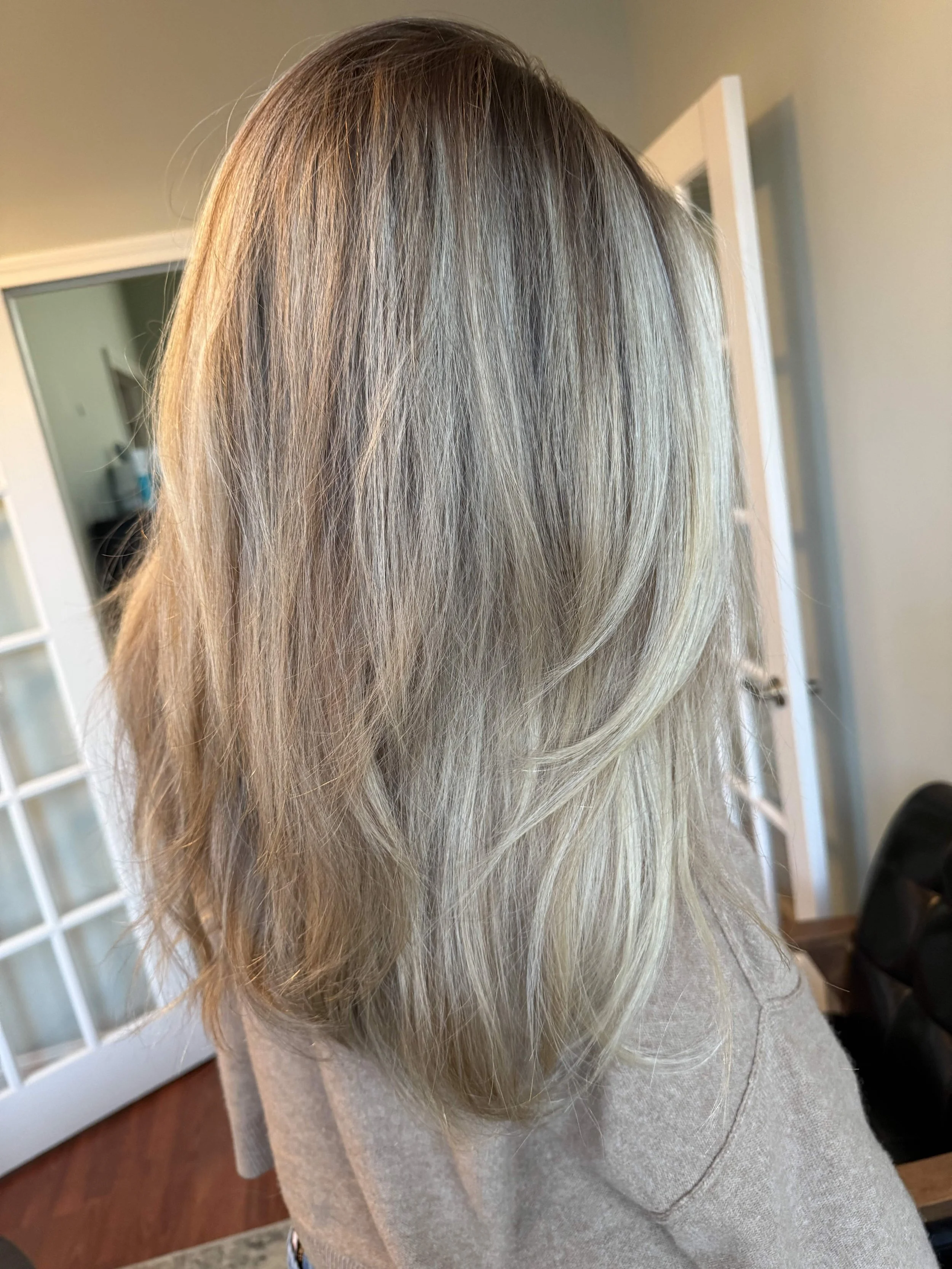 Woman with shoulder-length blonde hair, styled with layers, indoors near a door, wearing a beige top.