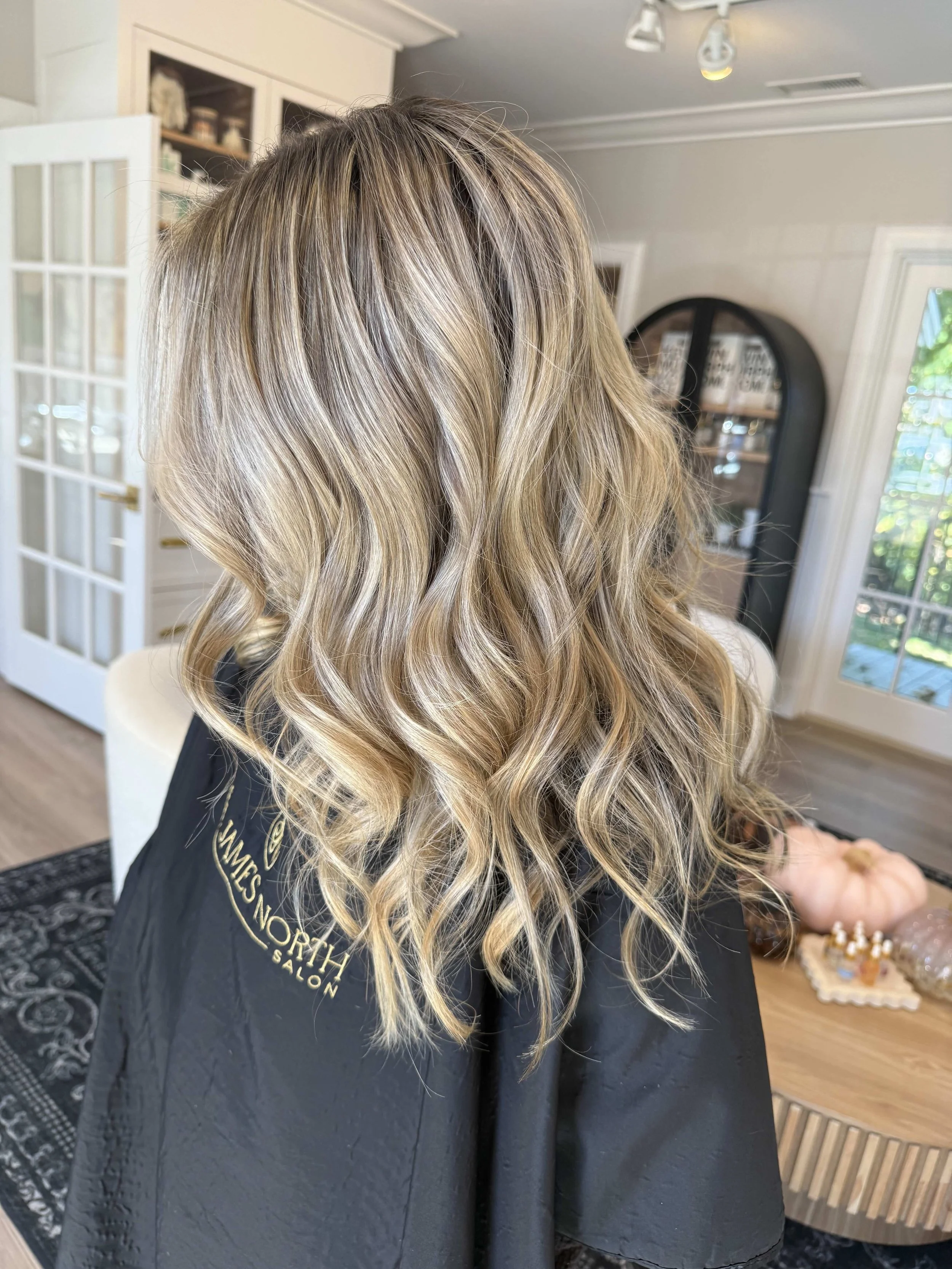 Blonde woman with wavy hair sitting in a salon chair, wearing a black salon cape, in a room with white walls and glass doors.