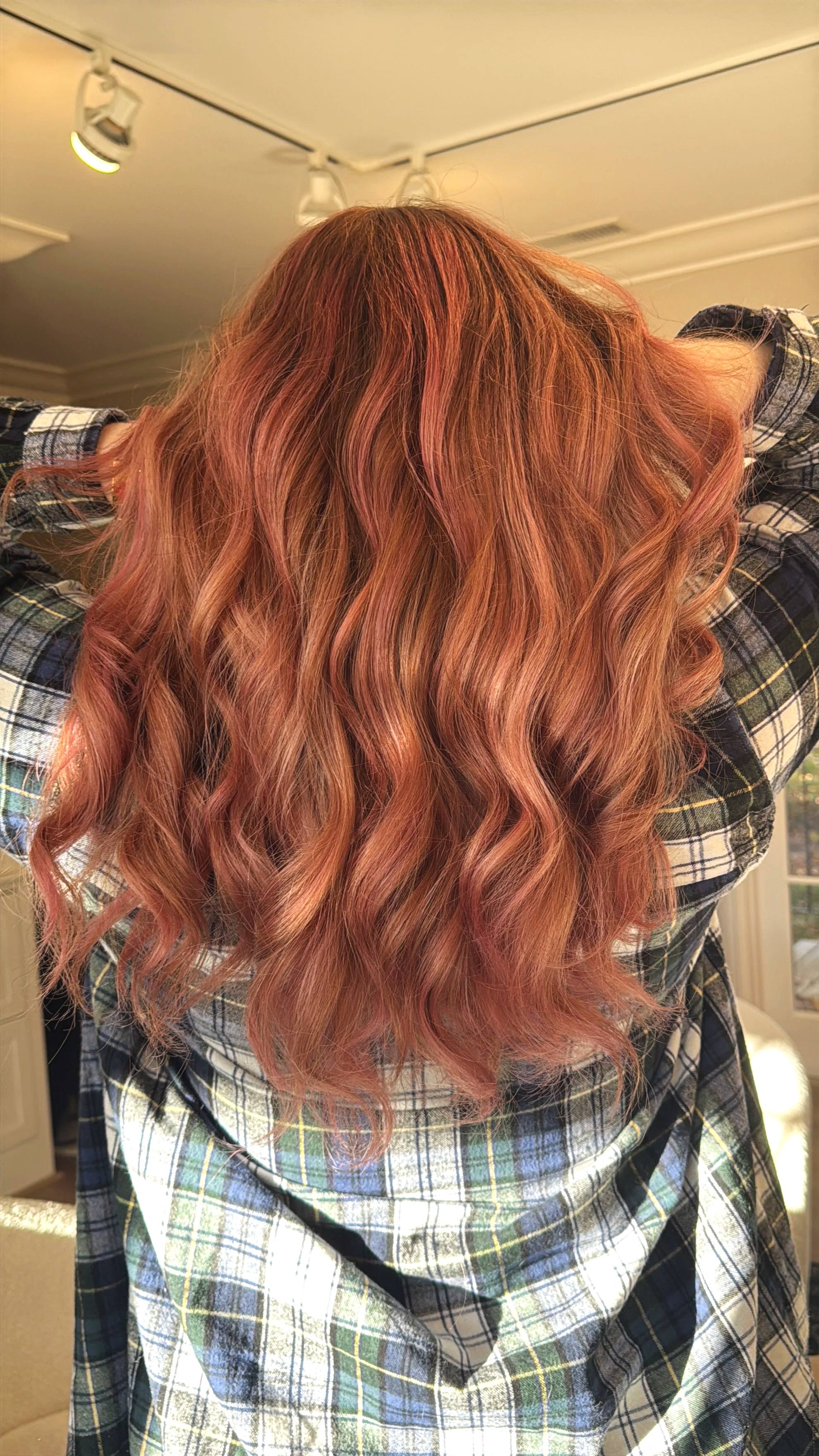 Person with long wavy red hair seen from the back, wearing a plaid shirt indoors.