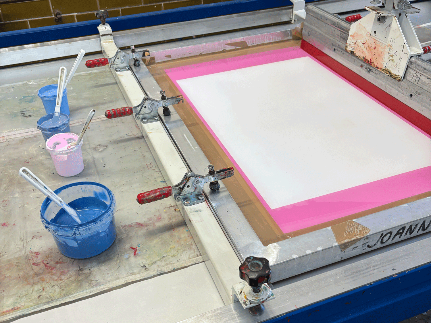 Screen printing frame with a mostly blank screen, ink containers with blue, pink, and purple inks, and squeegees on a work surface.