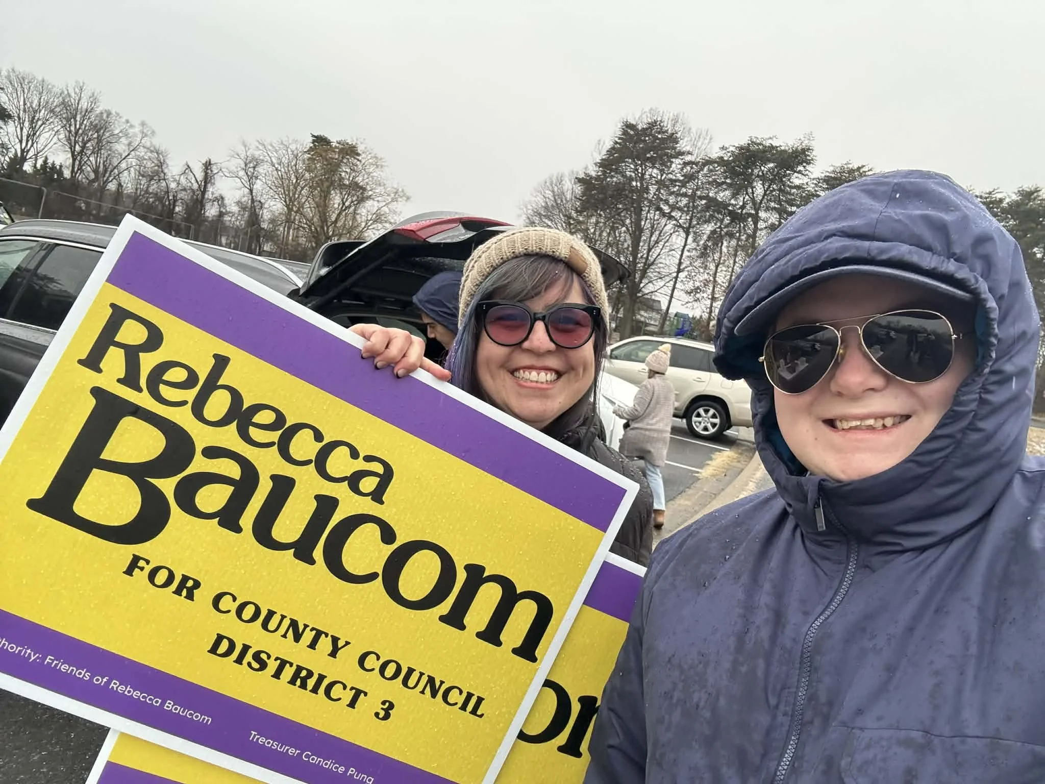 Two people smiling and holding a campaign sign for Rebecca Baucom, running for County Council District 3, in a parking lot on a rainy day. One woman wears sunglasses, a blue raincoat, and a hood; the other wears a beige knit hat and sunglasses.