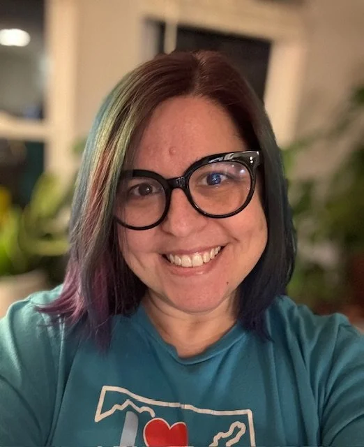 A woman smiling with glasses and multicolored hair in an indoor setting with houseplants in the background.