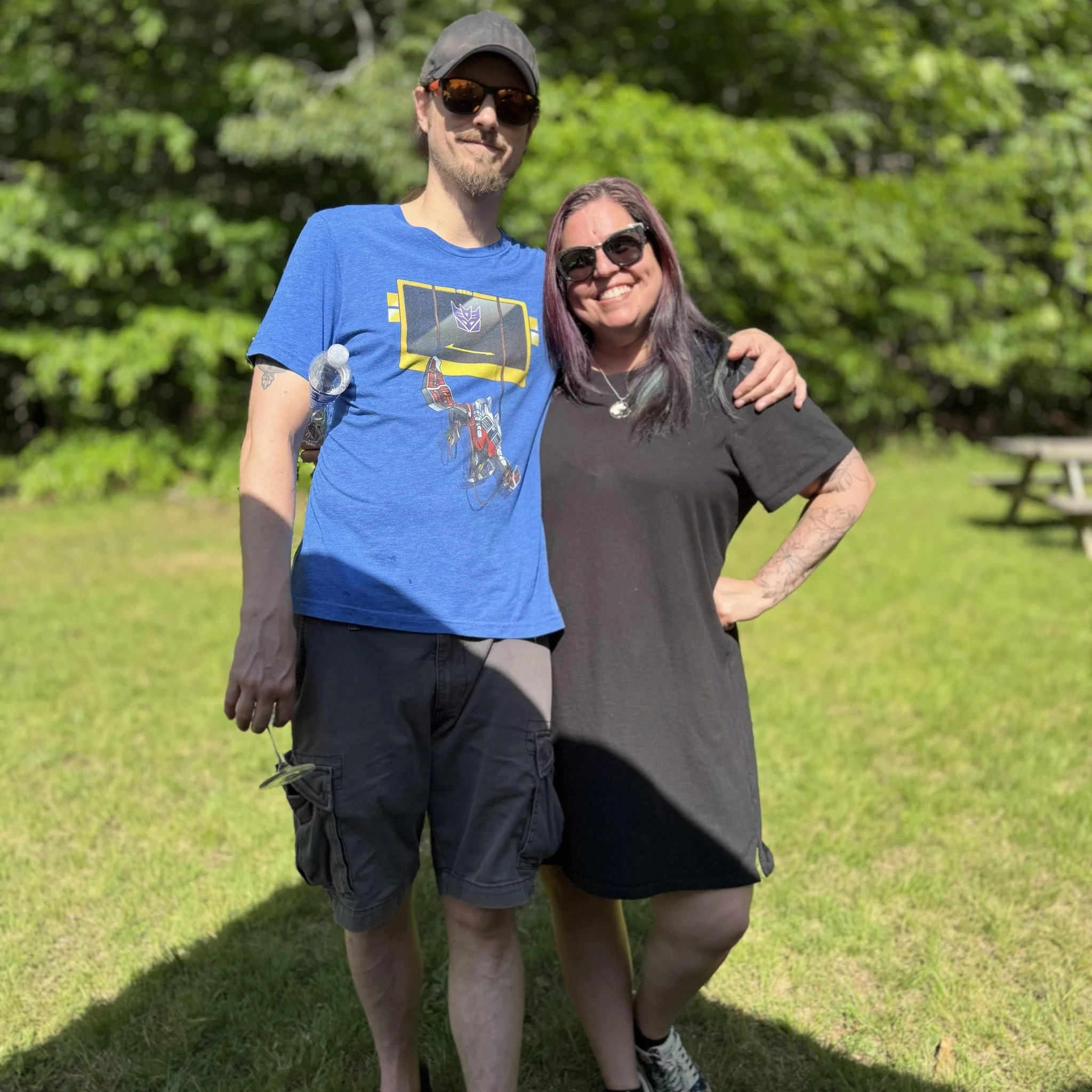 A man and a woman standing outdoors on a grassy area, smiling and posing with their arms around each other, with trees in the background.