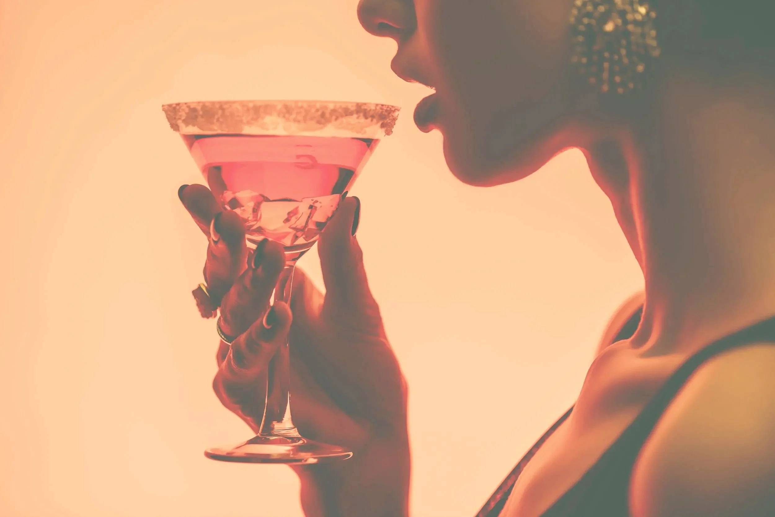 Close-up of a woman holding a pink cocktail glass with a salted rim, about to take a sip, background is warm and softly lit.
