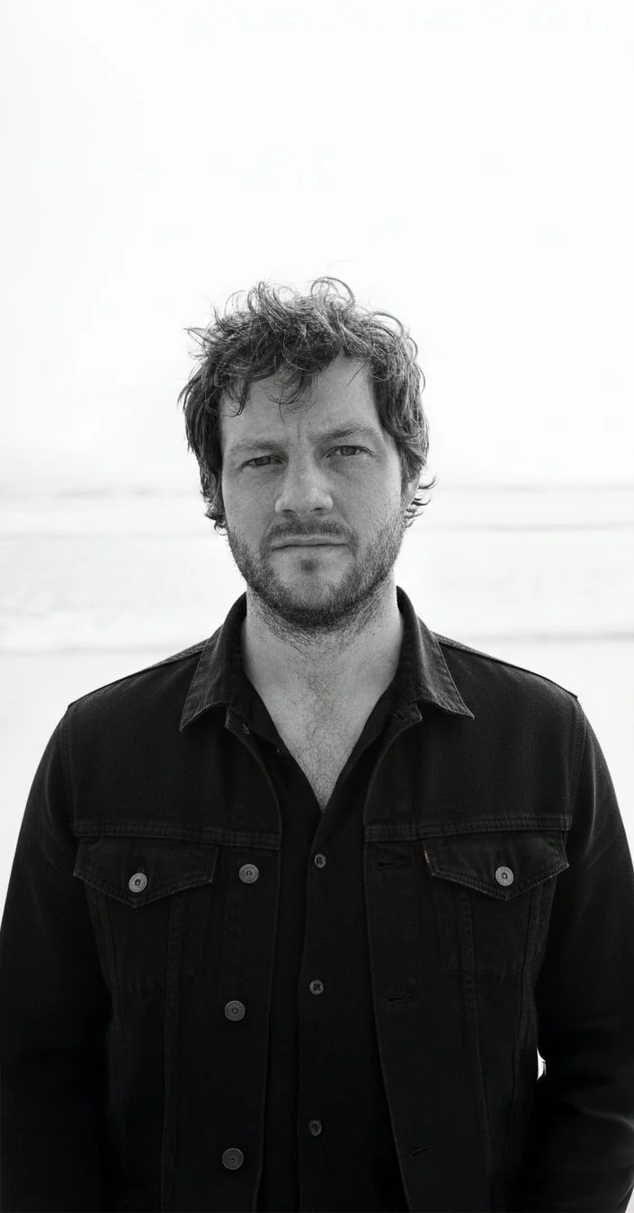 Black and white photo of a man with wavy hair and beard, wearing a black jacket, standing outdoors with a beach and ocean in the background.