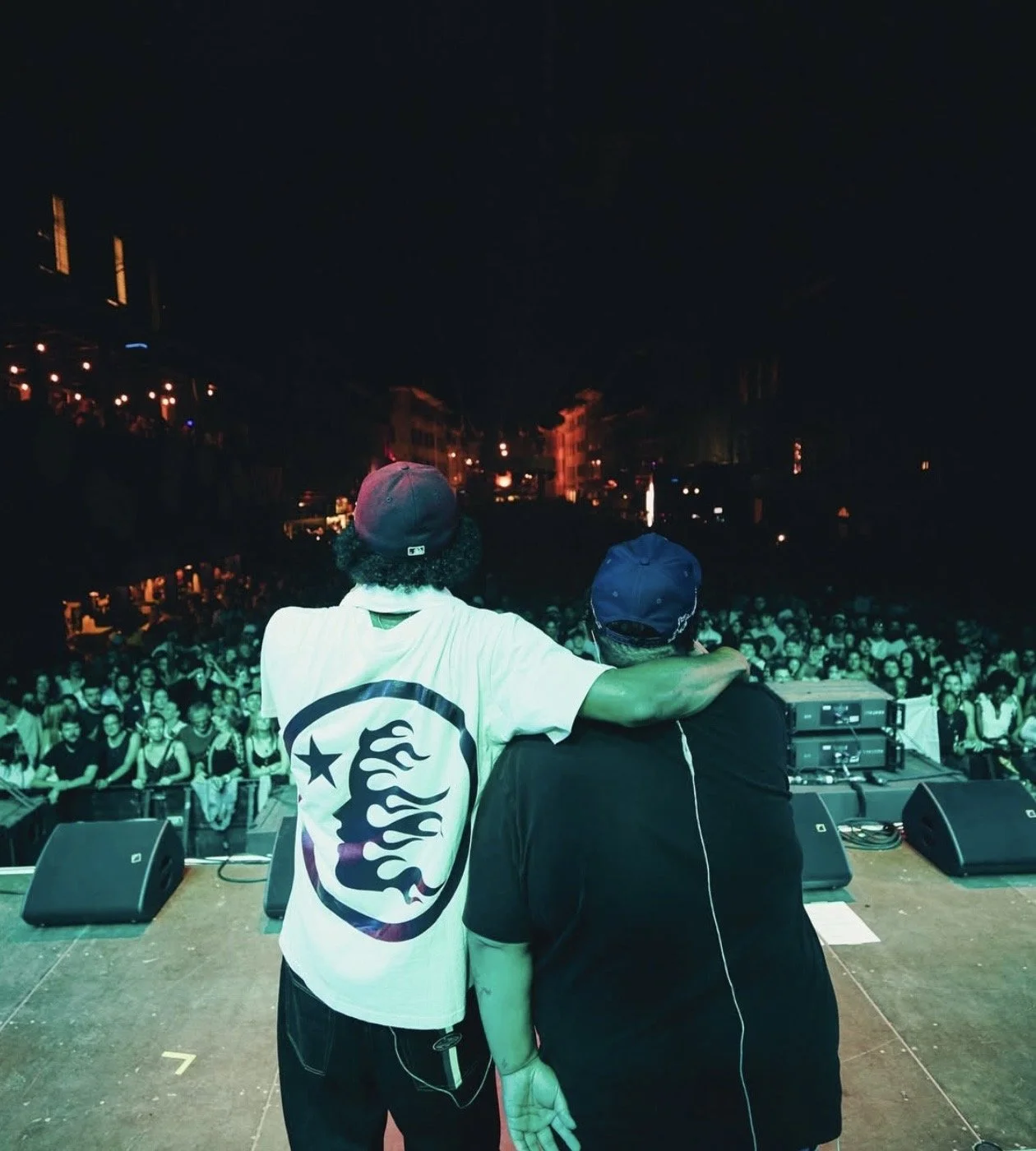 Zwei Männer auf einer Bühne bei einem Konzert oder Festival, einer mit Arm um den anderen, im Hintergrund ein großes Publikum und Lichter in der Nacht.