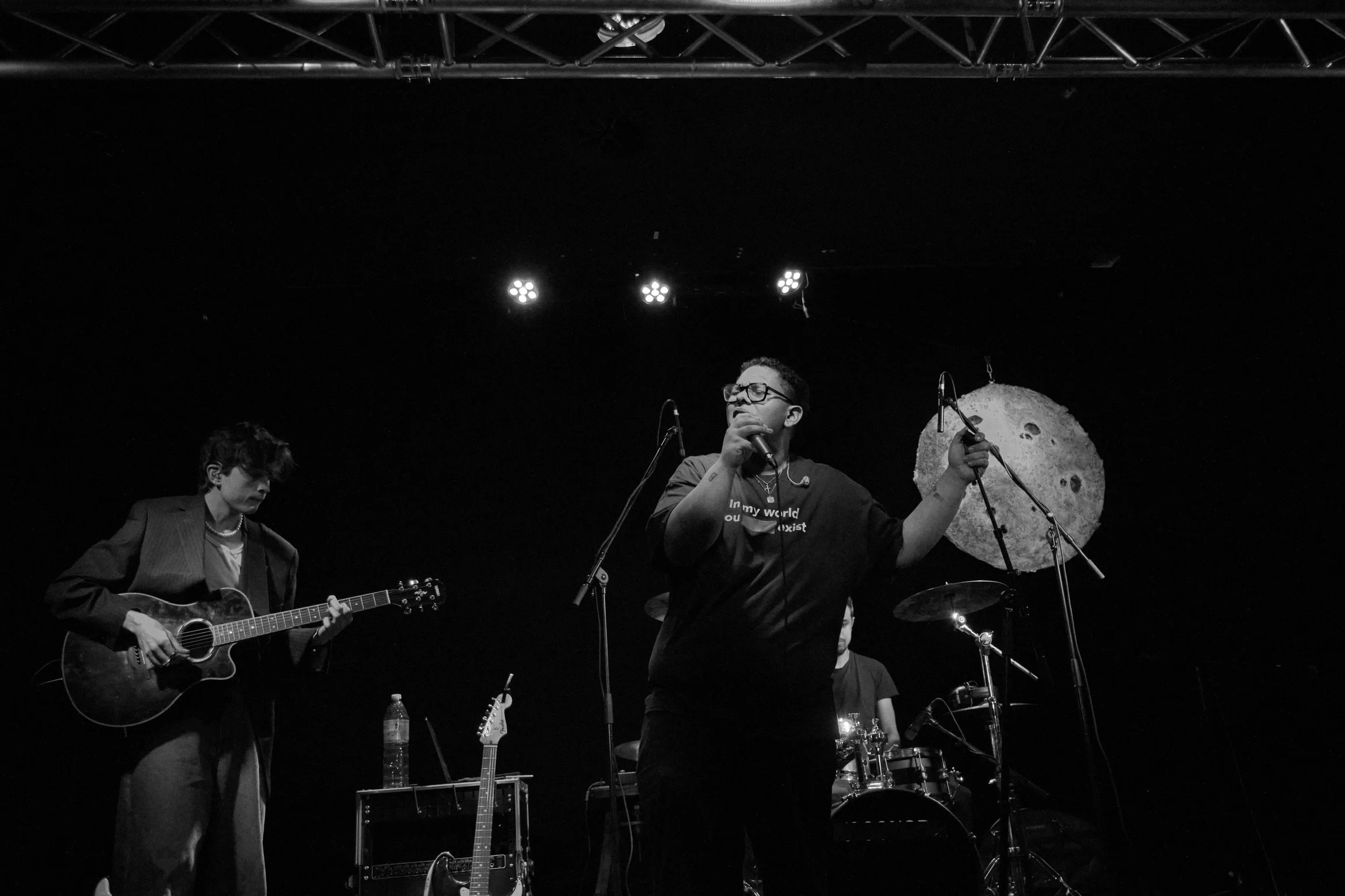 Eine Band spielt auf einer Bühne, die Sängerin hält ein Mikrofon und singt, während die Gitarristin neben ihr Gitarre spielt. Im Hintergrund ist der Schlagzeuger. Es gibt eine große Mond-Dekoration an der Wand hinter der Gruppe.