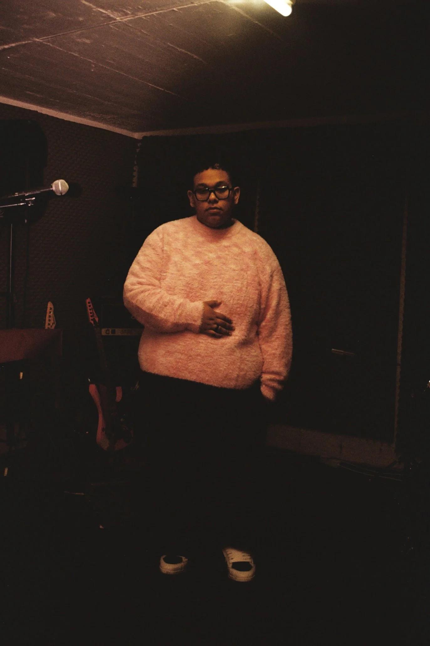 Junge Person in einem Tonstudio, trägt eine rosa Pullover, schwarze Brille, steht vor einem Mikrofon, umgeben von Musikinstrumenten und Aufnahmeausrüstung.