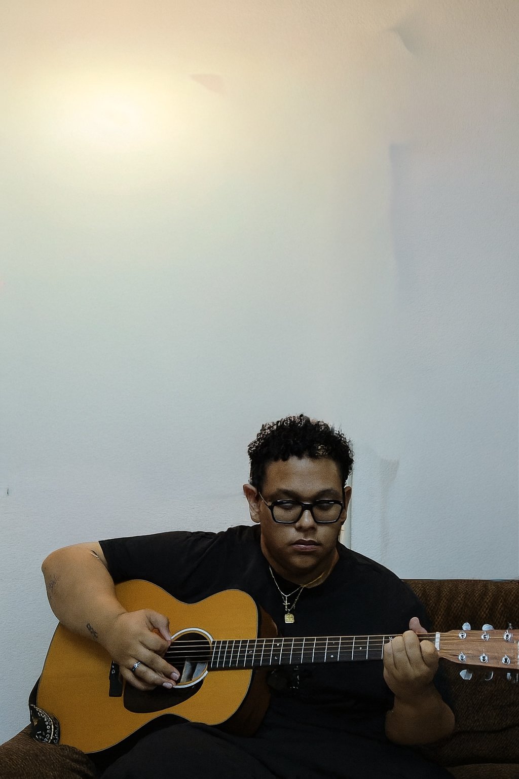 Junge Person mit Brille spielt Gitarre auf braunem Sofa in einem Raum mit weißer Wand im Hintergrund.