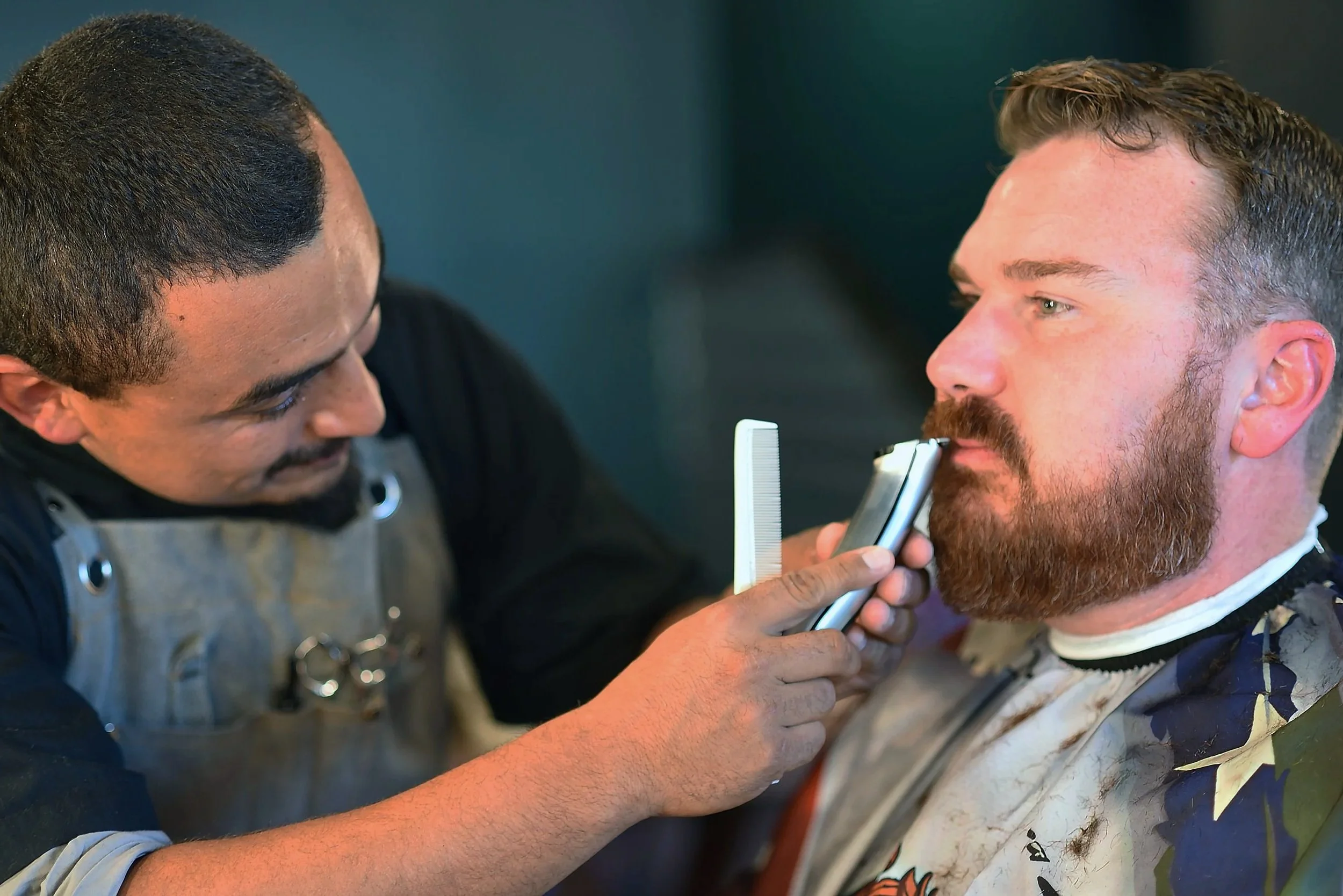 Barber giving a beard trim with electric trimmer to a male customer in a barbershop.
