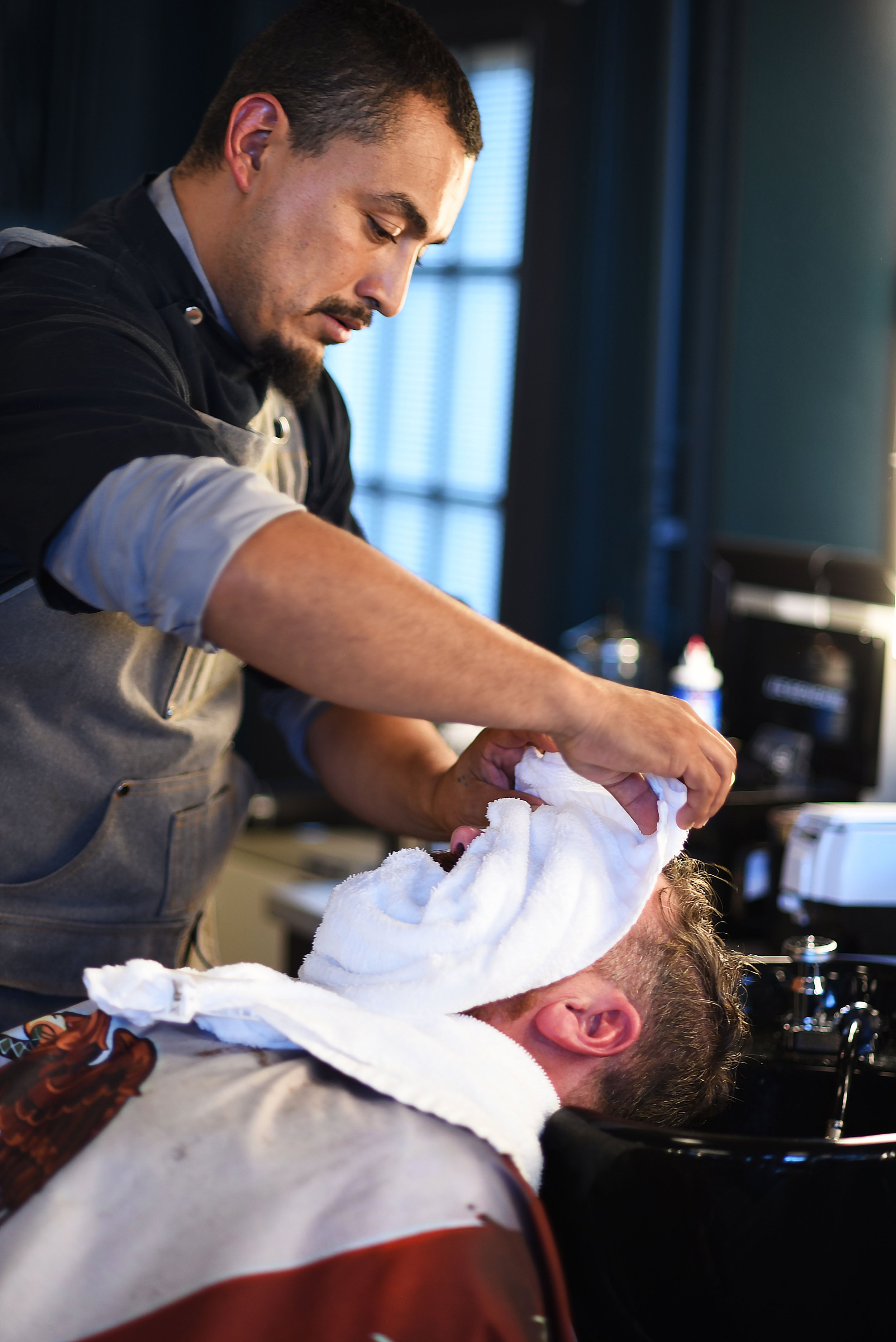 Barber wrapping a hot towel around a man's face with the man reclined and relaxed, and the barber focused on his work.
