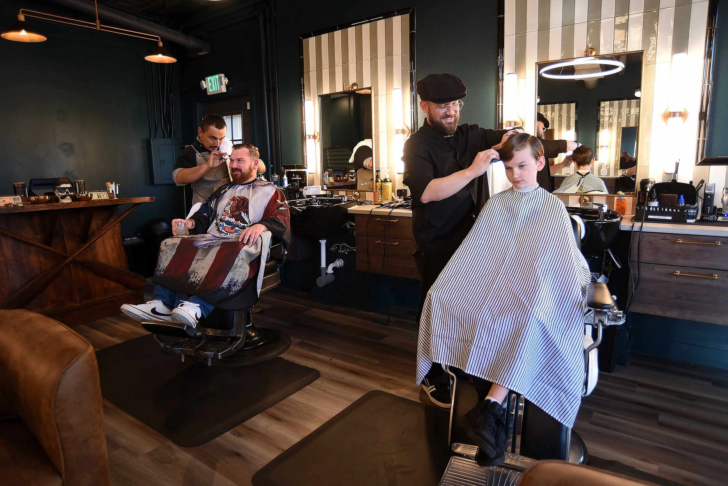 Barber giving a young boy a haircut in a modern barbershop, with another customer seated nearby.
